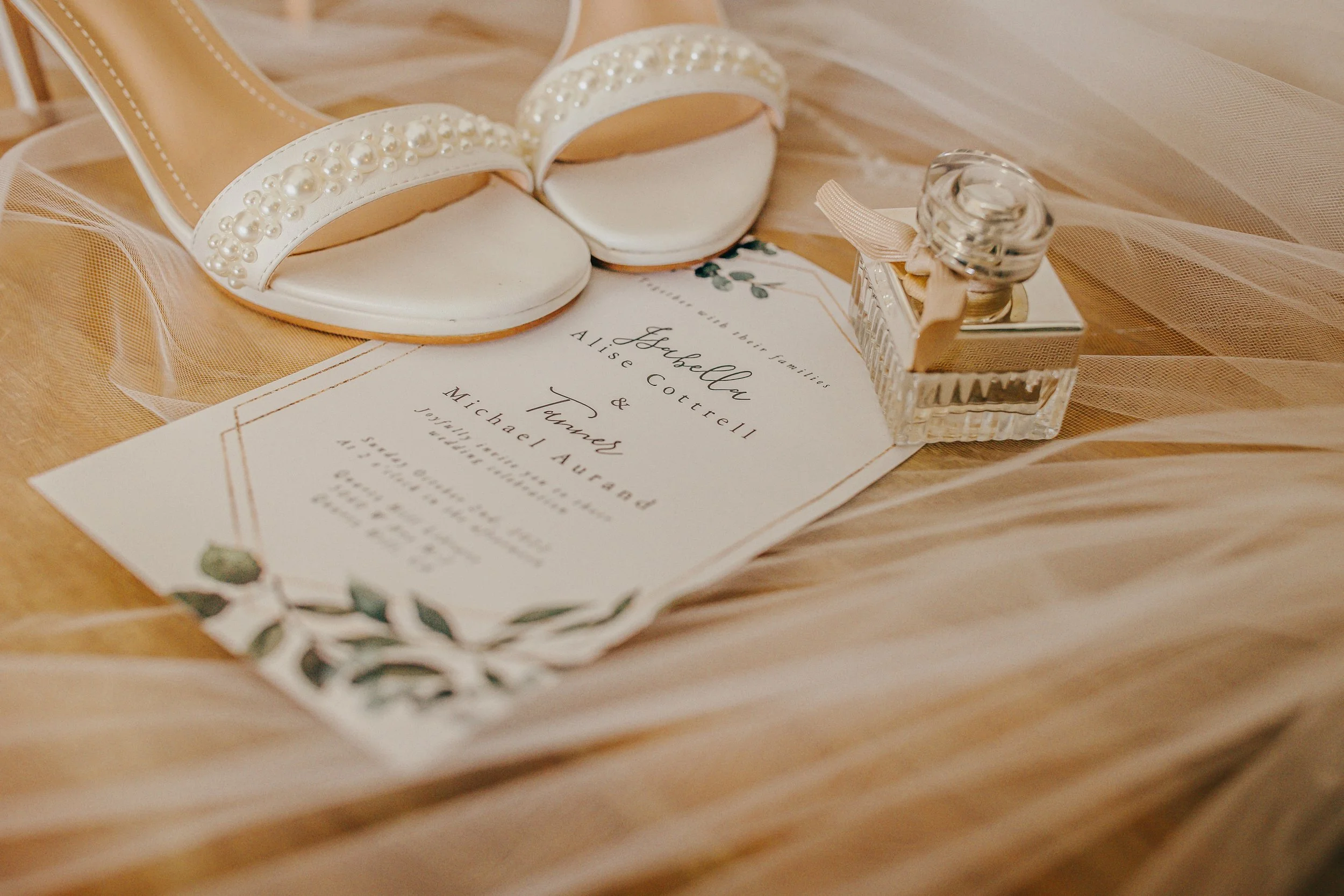 A pair of white wedding shoes with pearl embellishments, a wedding invitation with green leaf illustrations, and a small glass bottle tied with a ribbon, all placed on a soft fabric surface.