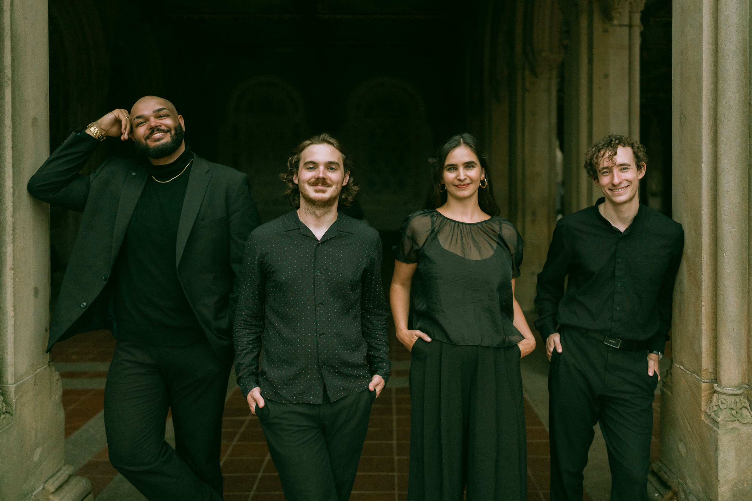Four diverse people standing in a stone archway, smiling at the camera, dressed in black clothing, in a historical or architectural setting.