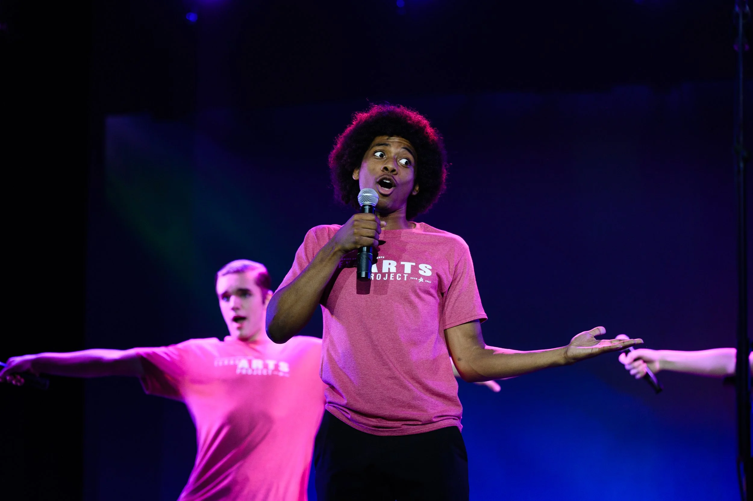A young person with curly hair singing into a microphone on stage, wearing a pink t-shirt with the words 'ARTS PROJECT' printed on it, with two other performers in pink shirts seemingly dancing or gesturing behind them.