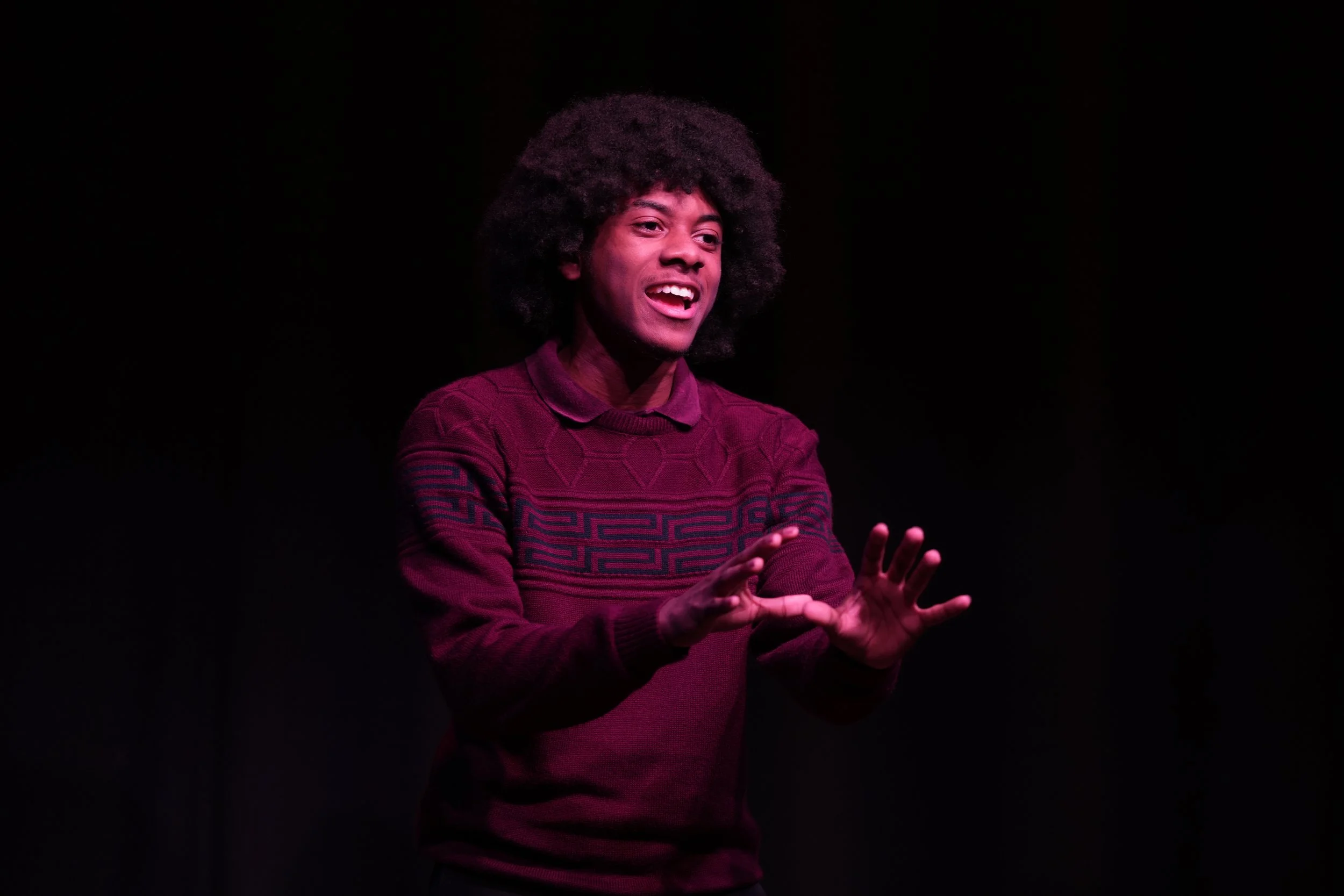 A young man with curly hair wearing a maroon sweater is speaking on stage with a dark background.