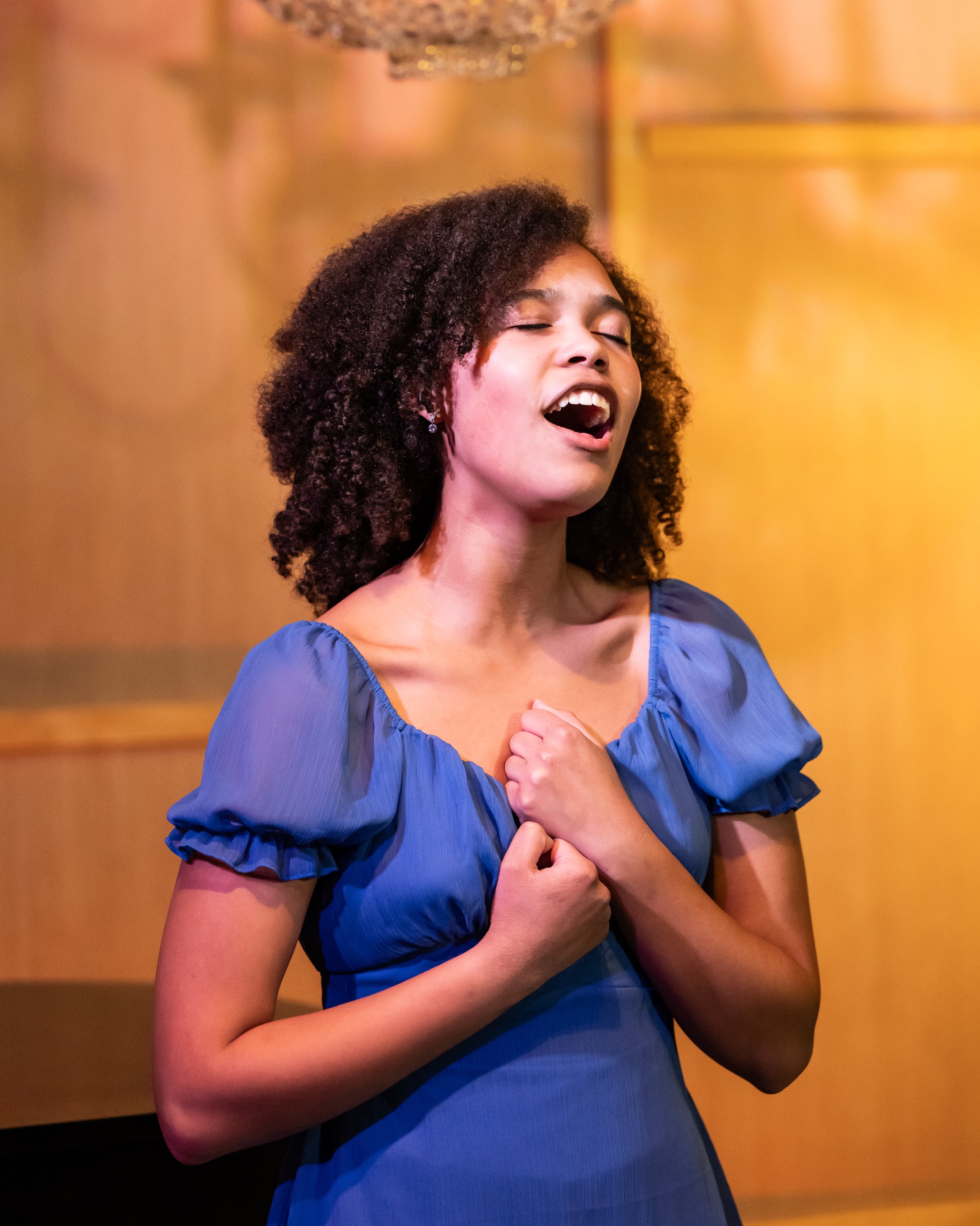A woman with curly hair and a blue dress singing with her eyes closed and hands clasped near her chest, standing in front of a golden background.