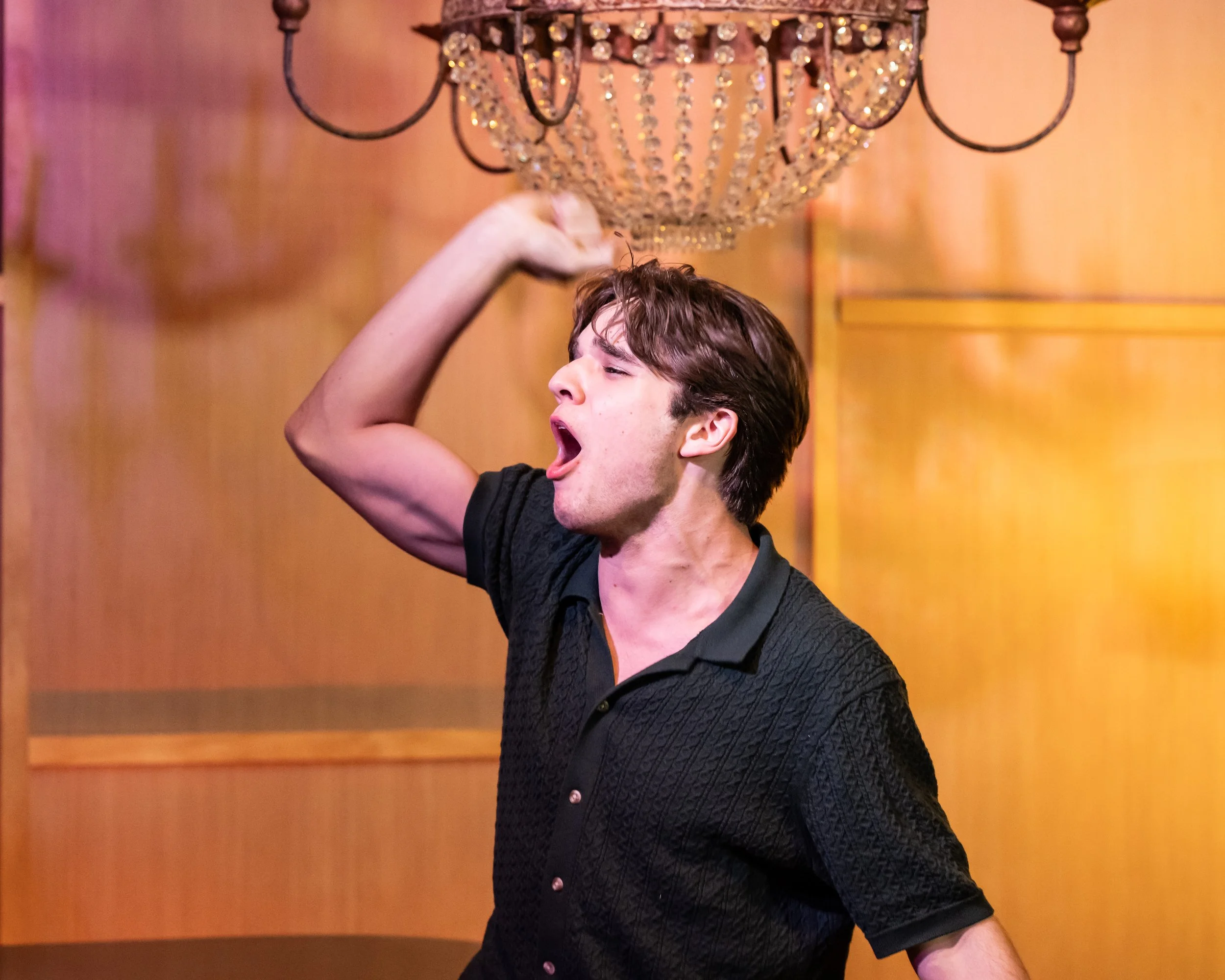 A young man with brown hair and wearing a black shirt is yawning with his mouth open, standing indoors under a chandelier.