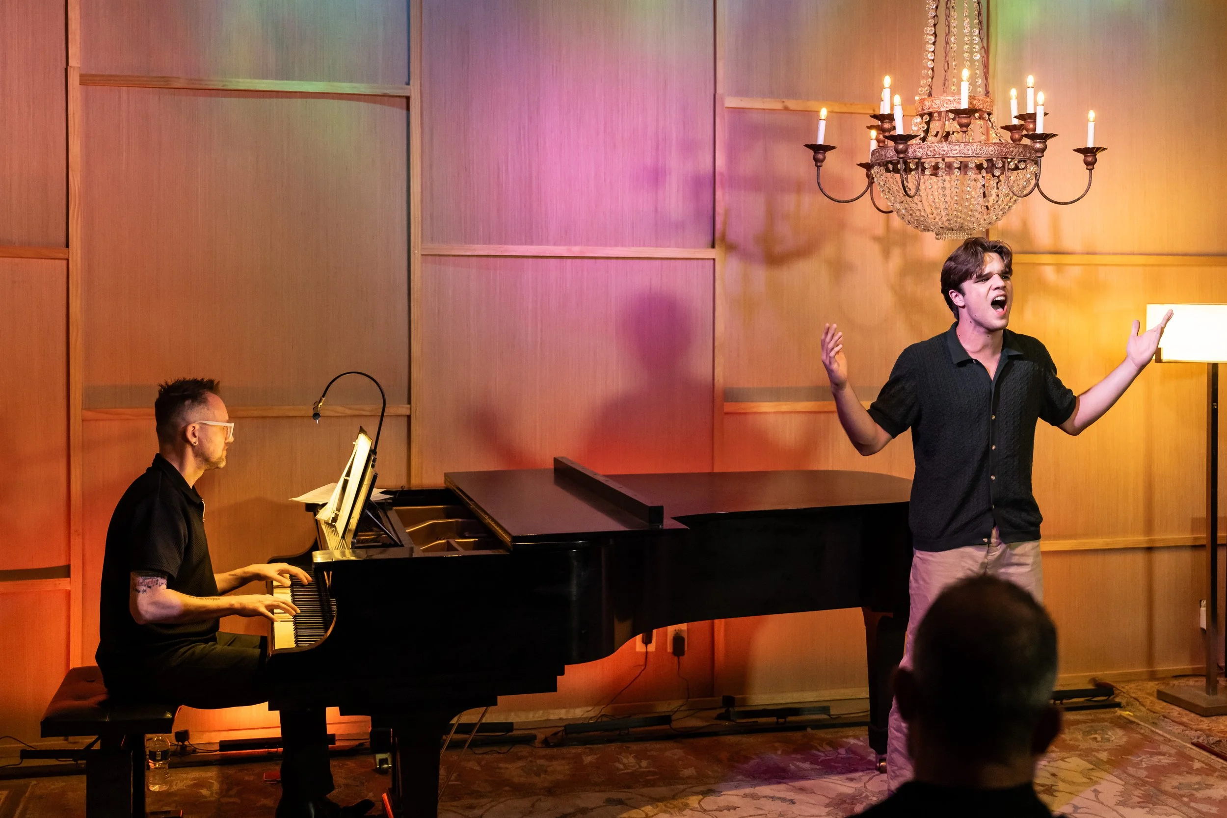 A man singing passionately on stage with a pianist playing on a grand piano beside him, in a room with warm lighting and a decorative chandelier hanging from the ceiling.