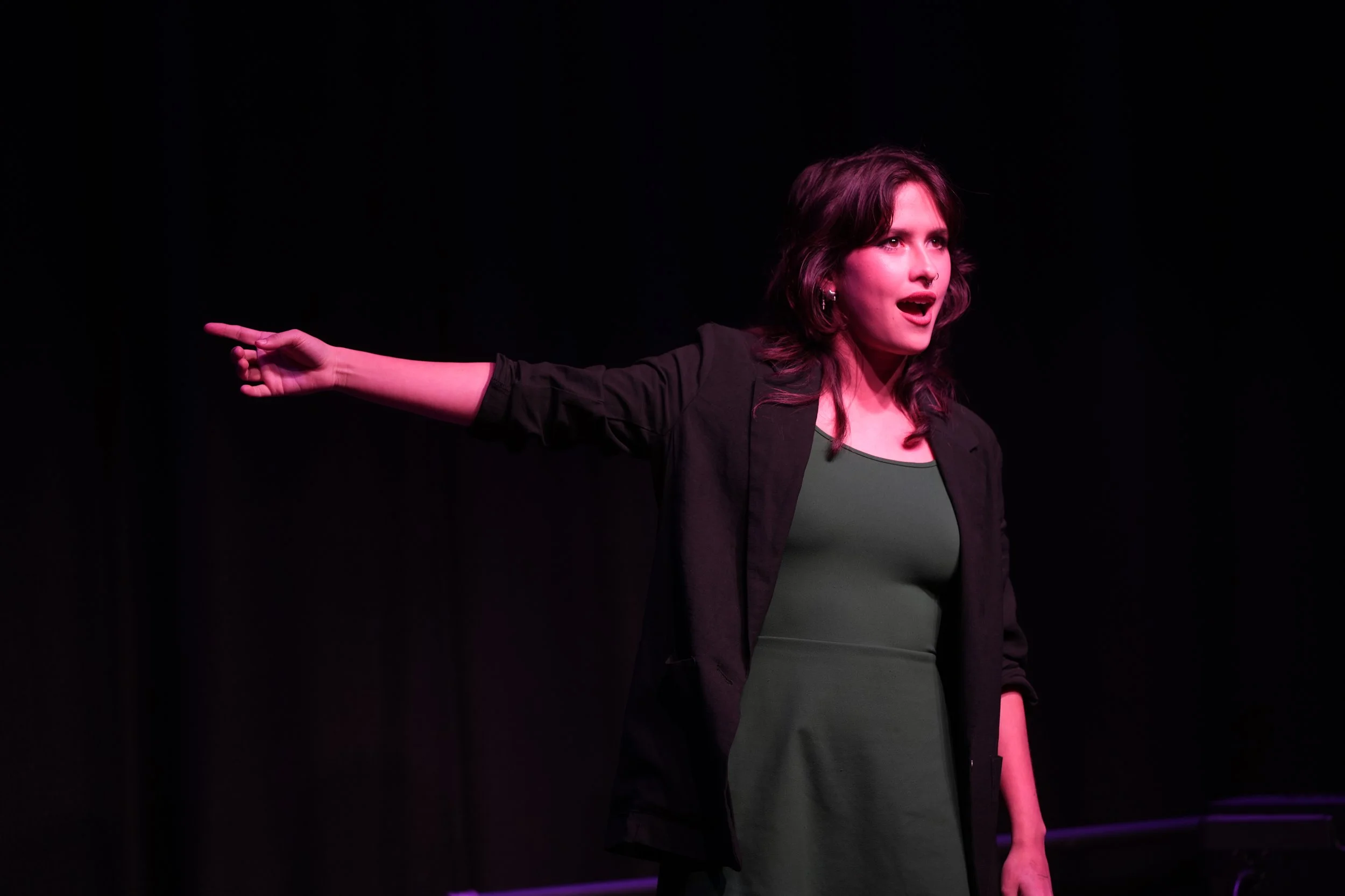 A woman with shoulder-length dark hair, wearing a black jacket and green dress, is standing on stage with one arm extended to the side, pointing, and appears to be speaking or acting with an expressive facial expression.