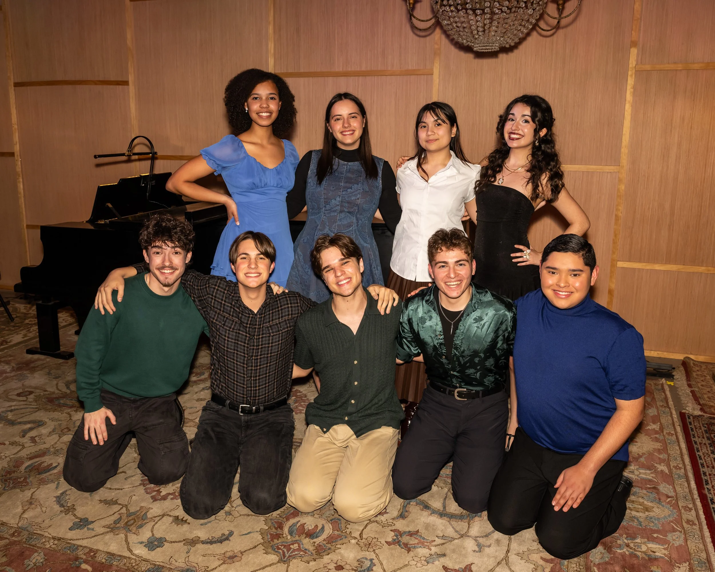 Group of ten young adults posing together in a music studio with a grand piano and wooden panels, smiling and dressed in casual and semi-formal clothing.