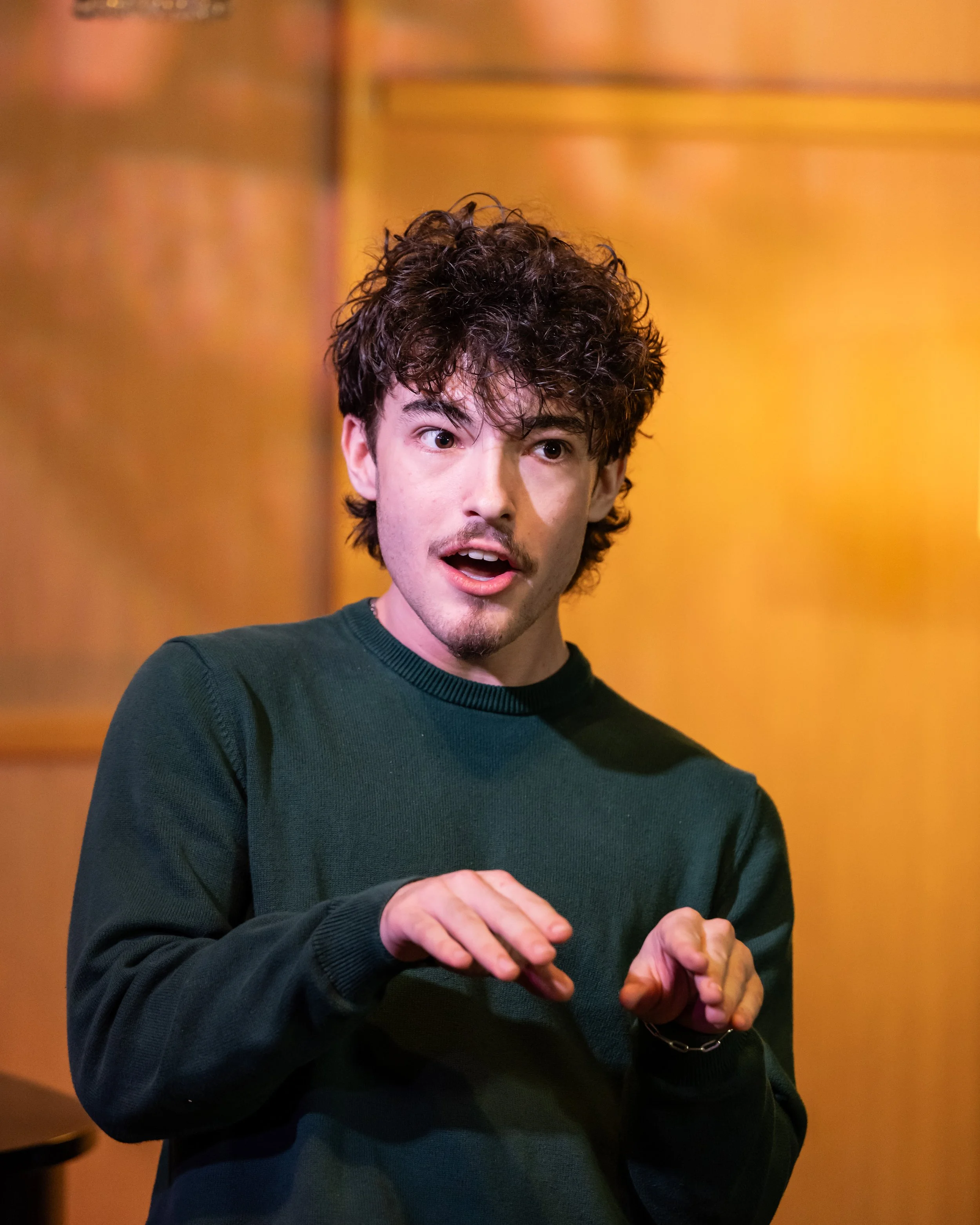 A young man with curly brown hair and a light beard, wearing a dark green sweater, is gesturing with his hands and speaking in an indoor setting with warm yellow walls.