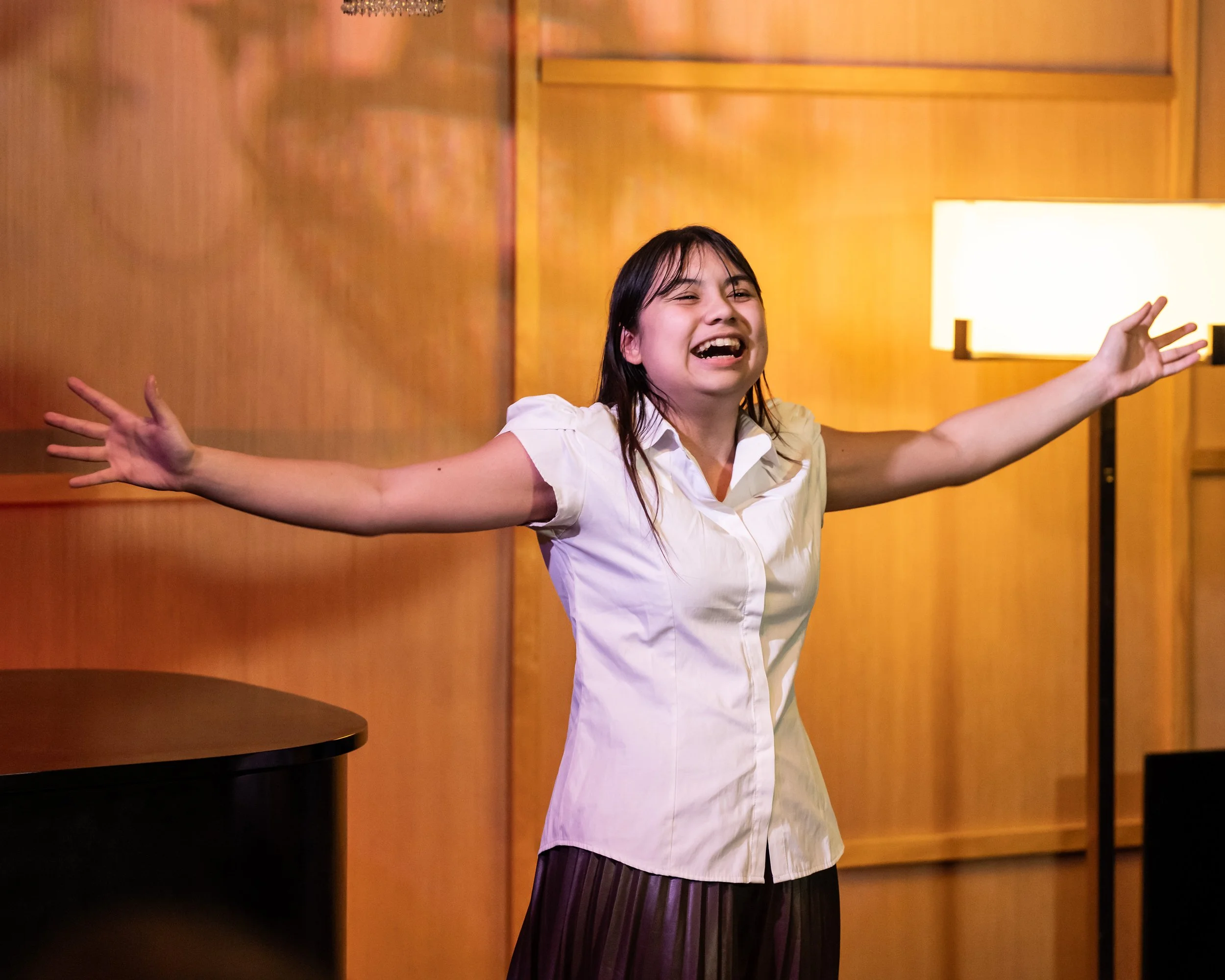 A woman with long dark hair, wearing a white shirt, is smiling widely with her arms outstretched in a warmly lit room with wooden paneling and a standing lamp.
