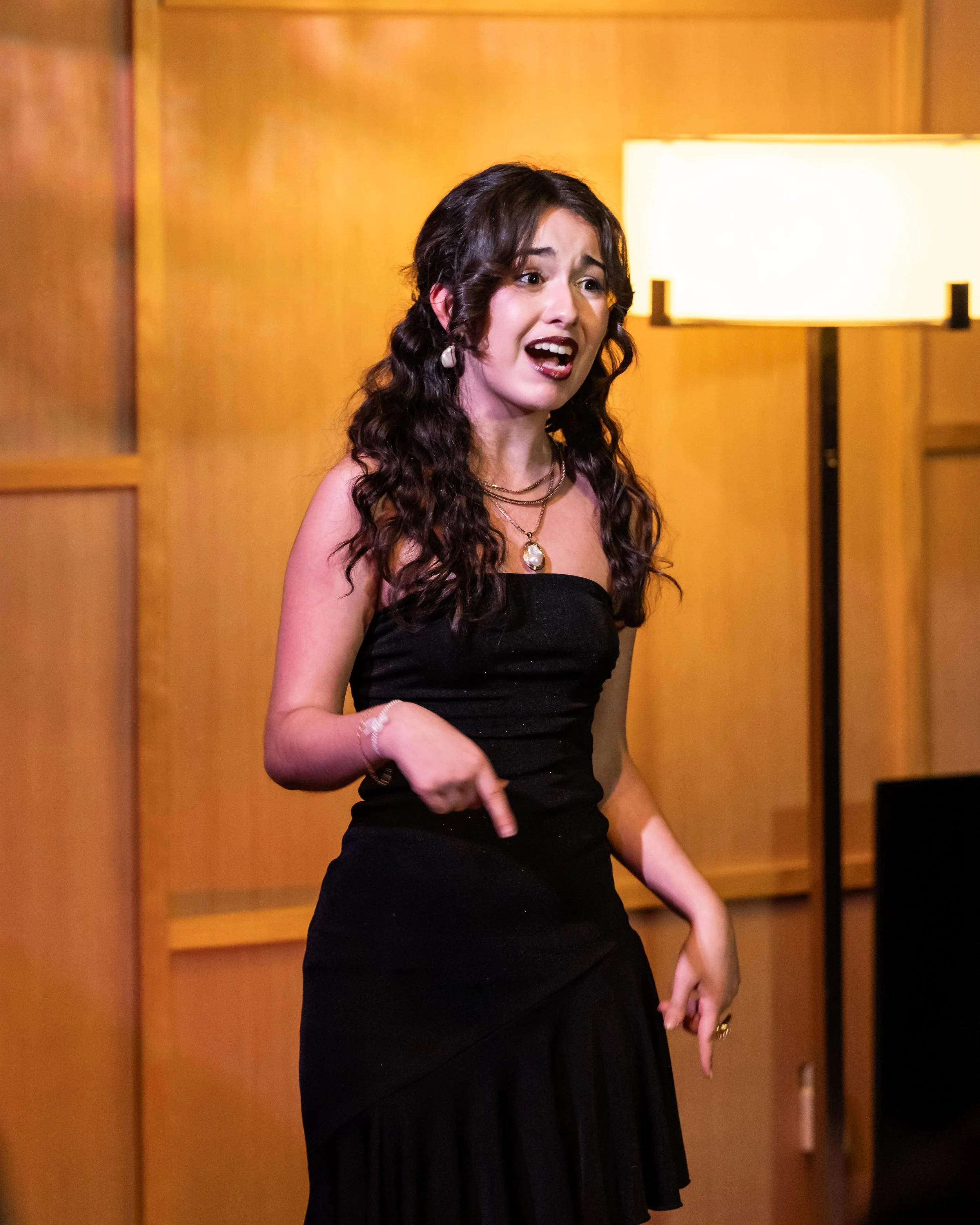A woman with curly dark hair, wearing a black strapless dress and layered necklaces, expresses surprise or shock in a room with warm wooden walls and a standing lamp.