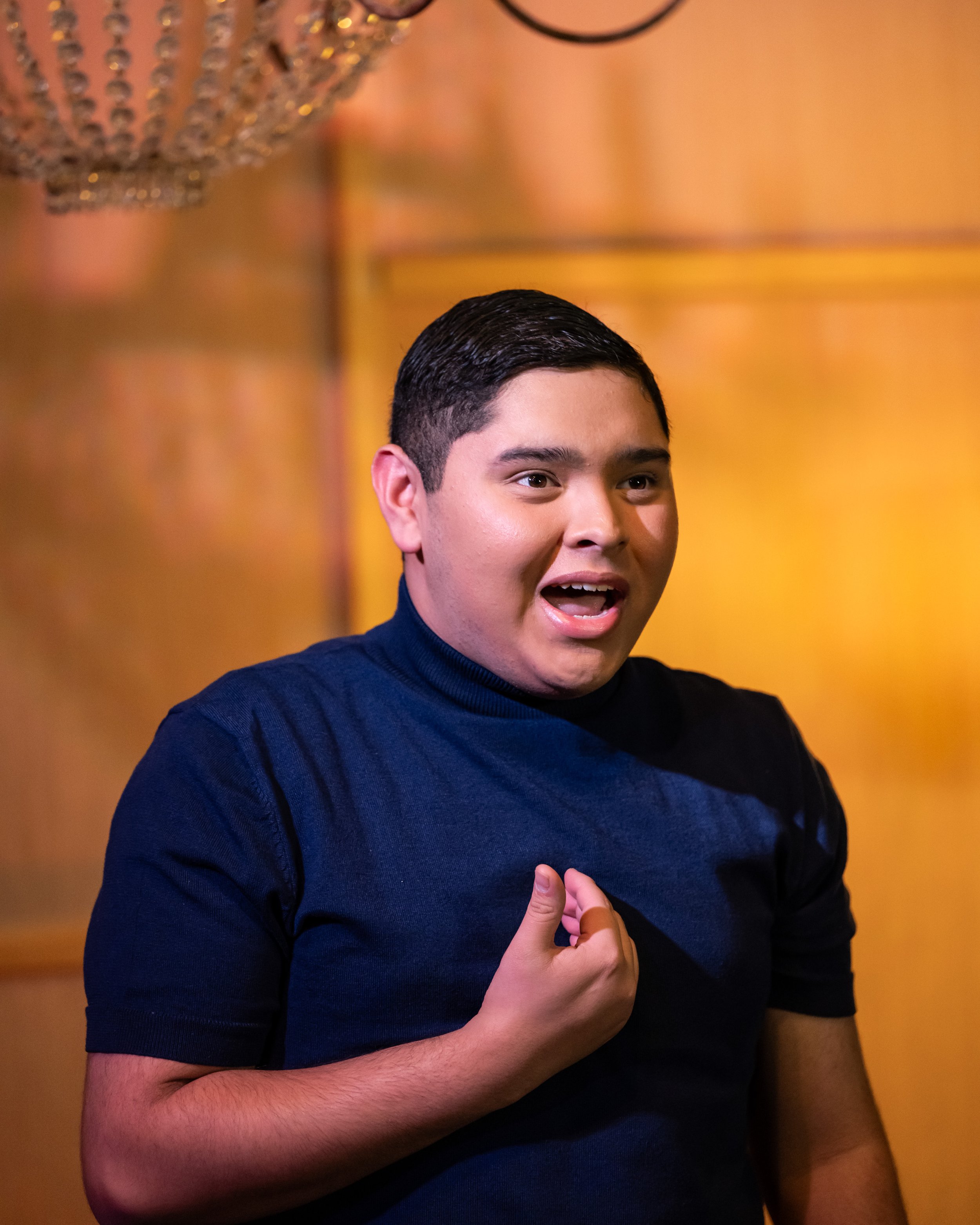 A young man in a navy blue shirt appears surprised or excited, with his mouth open and one hand over his chest, against a warm-colored background with a chandelier visible at the top.