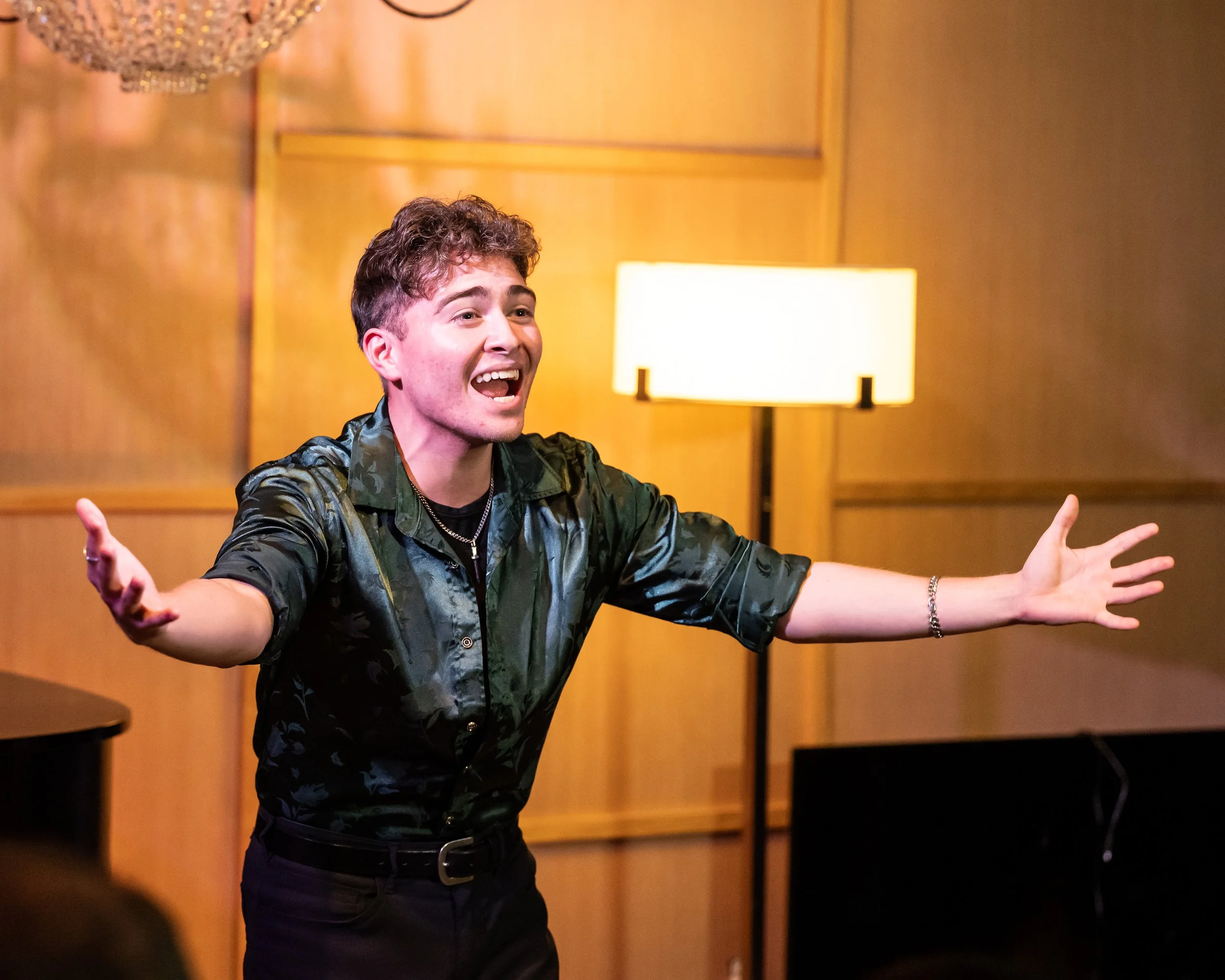 A young man with curly hair wearing a dark patterned shirt with rolled-up sleeves, standing with arms outstretched and smiling in a warmly lit room with wooden paneling and a standing lamp.