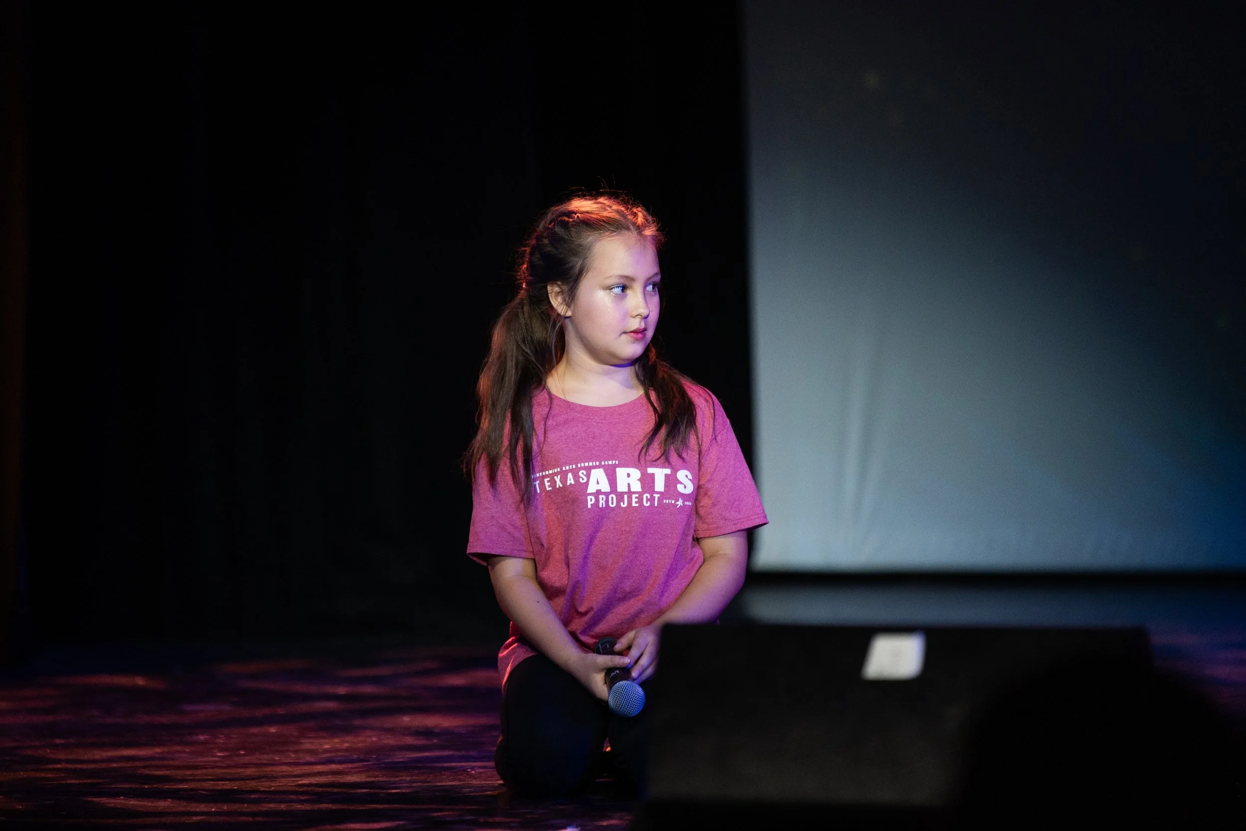 Young girl with long brown hair kneeling on stage, holding a microphone, wearing a pink T-shirt with 'TEXAS ARTS PROJECT' written on it, with a dark background and a large screen behind her.