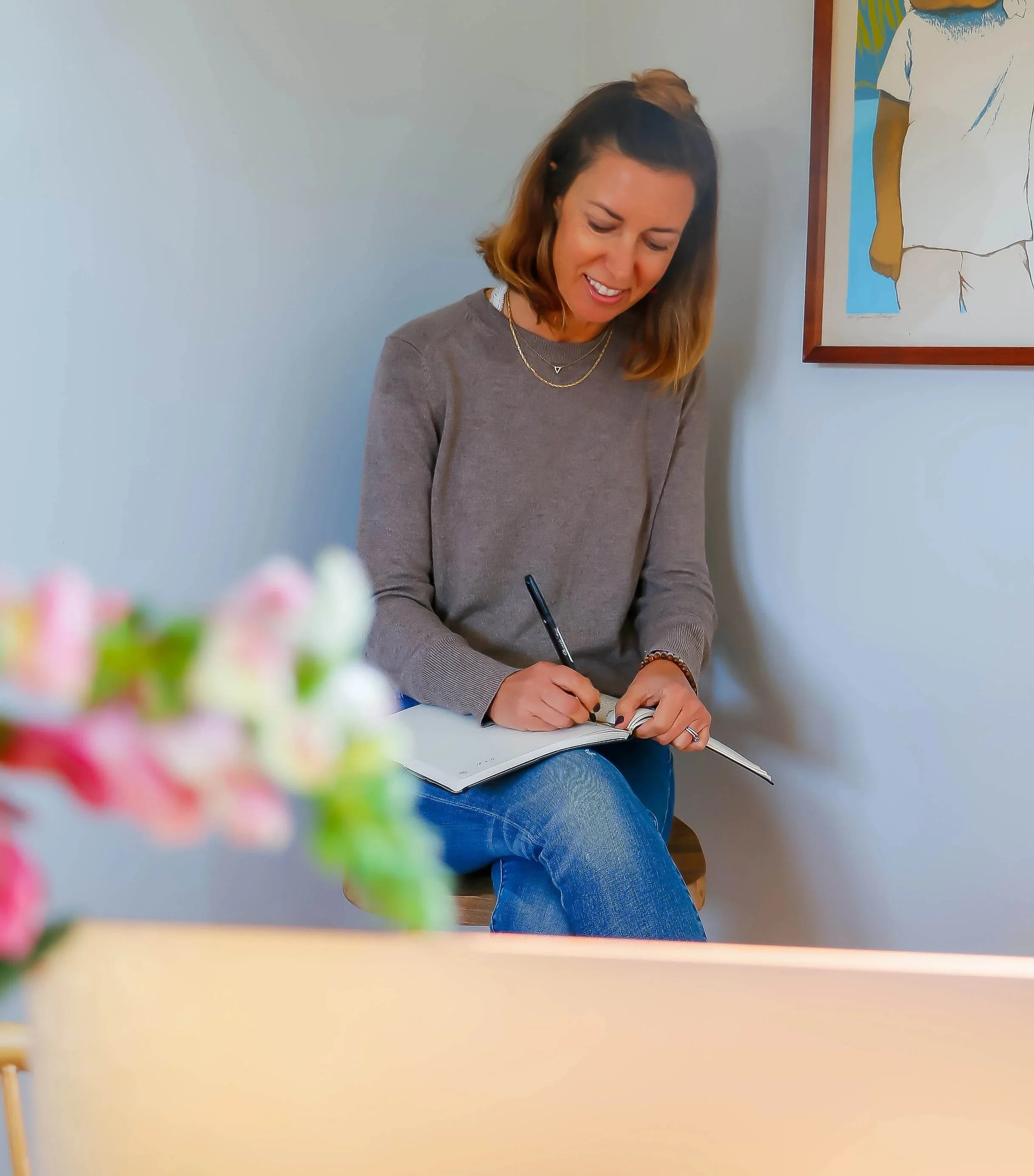 A woman with shoulder-length brown hair, wearing a gray sweater and blue jeans, sitting on a chair, smiling as she writes in a notebook.