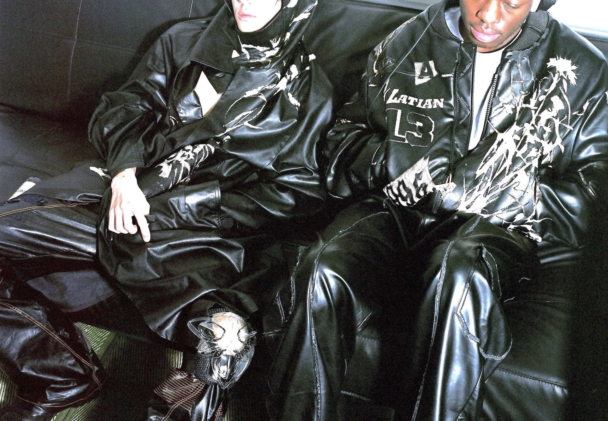 Two individuals sitting on a black leather couch, dressed in stylish black leather outfits, one featuring intricate designs and a "Latian 13" logo; the other with a draped scarf.