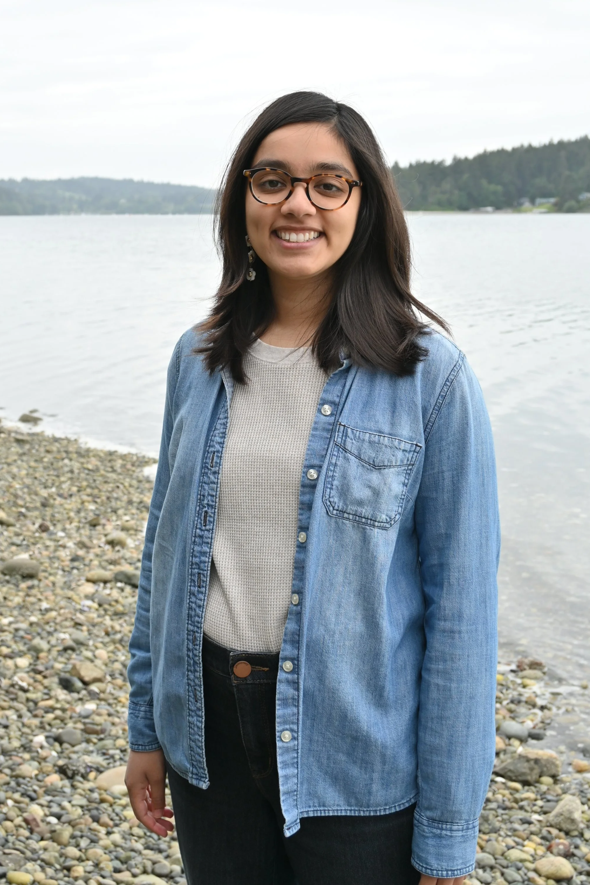 South Asian woman with shoulder length wavy black hair, glasses, and a blue buttondown over a white shirt stands on a rocky beach.