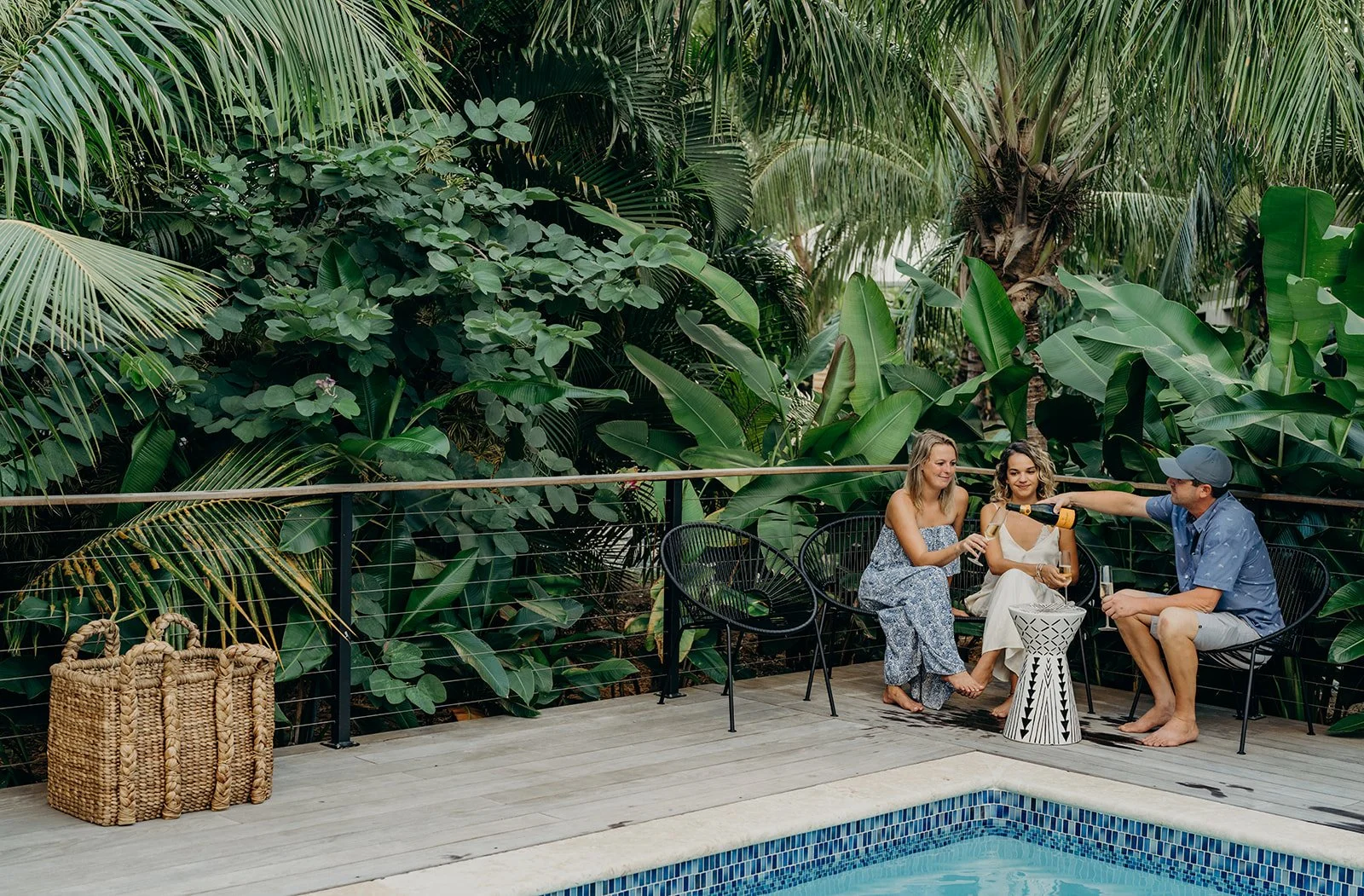 Three friends sitting beside a swimming pool, with lush green tropical plants in the background. One person is pouring champagne, and another is holding a glass, enjoying a moment together.