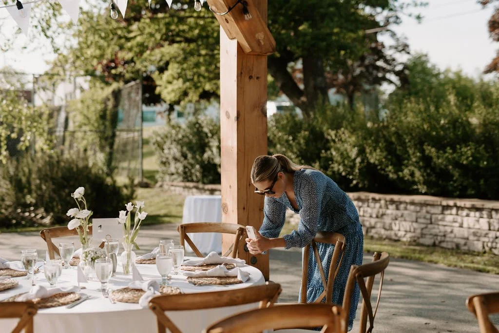 A woman in a blue dress takes a photo of a table set for an outdoor meal.