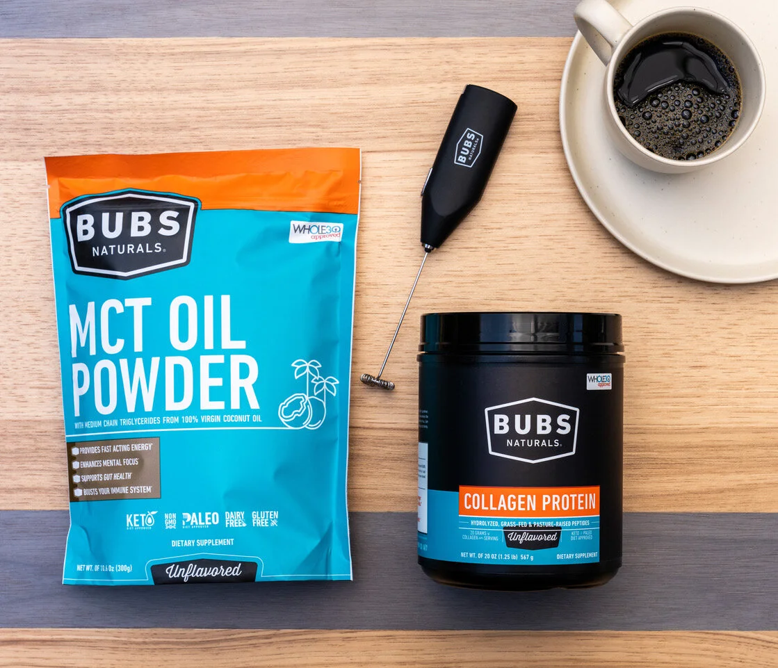 A top-down view of a wooden desk with a bag of BUBS Naturals MCT Oil Powder, a container of BUBS Naturals Collagen Protein, a coffee cup filled with black coffee on a saucer, and a small black tool with a thin metal lever.