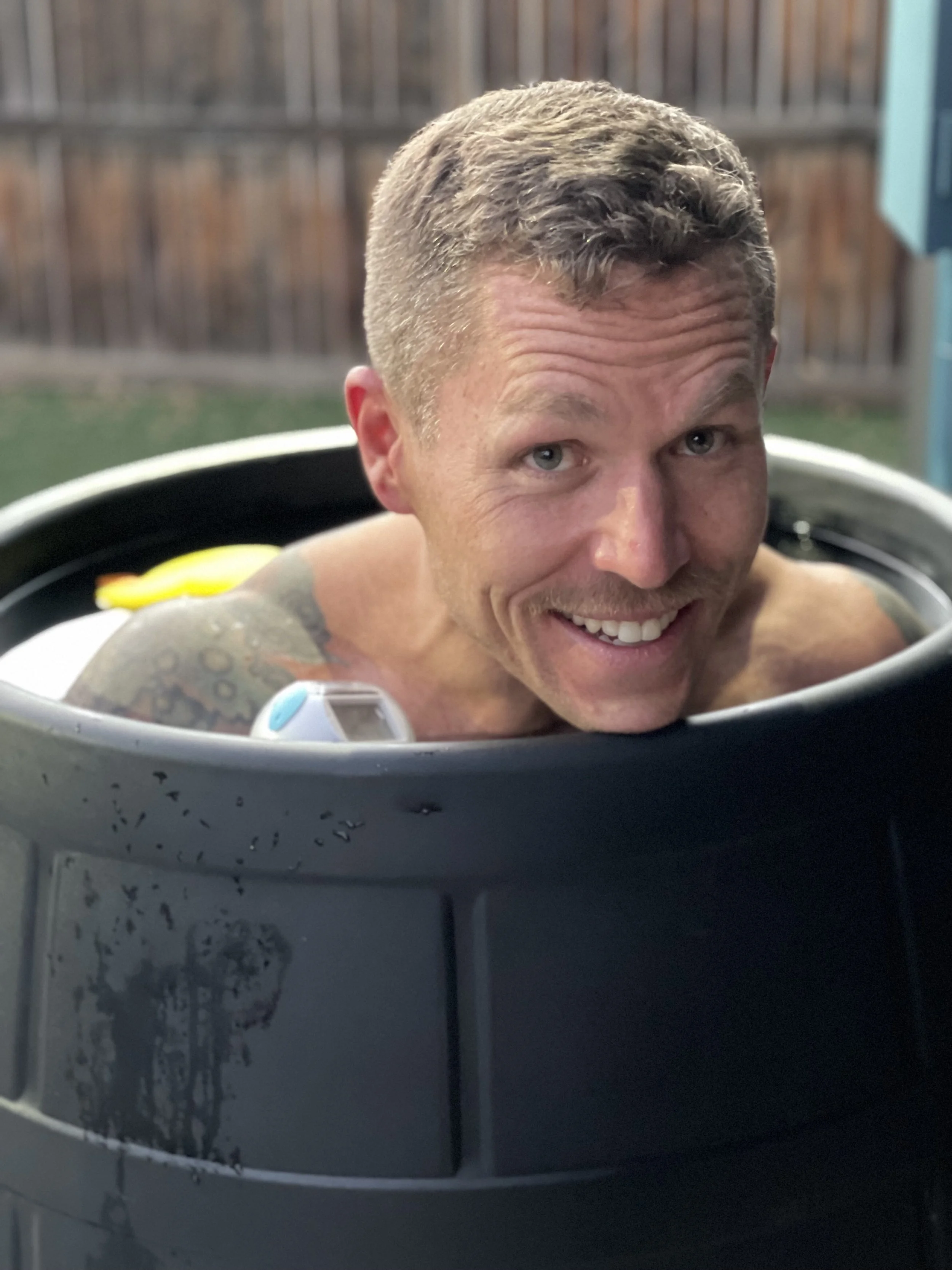Man smiling and looking over the edge of a black plastic barrel in an outdoor setting.