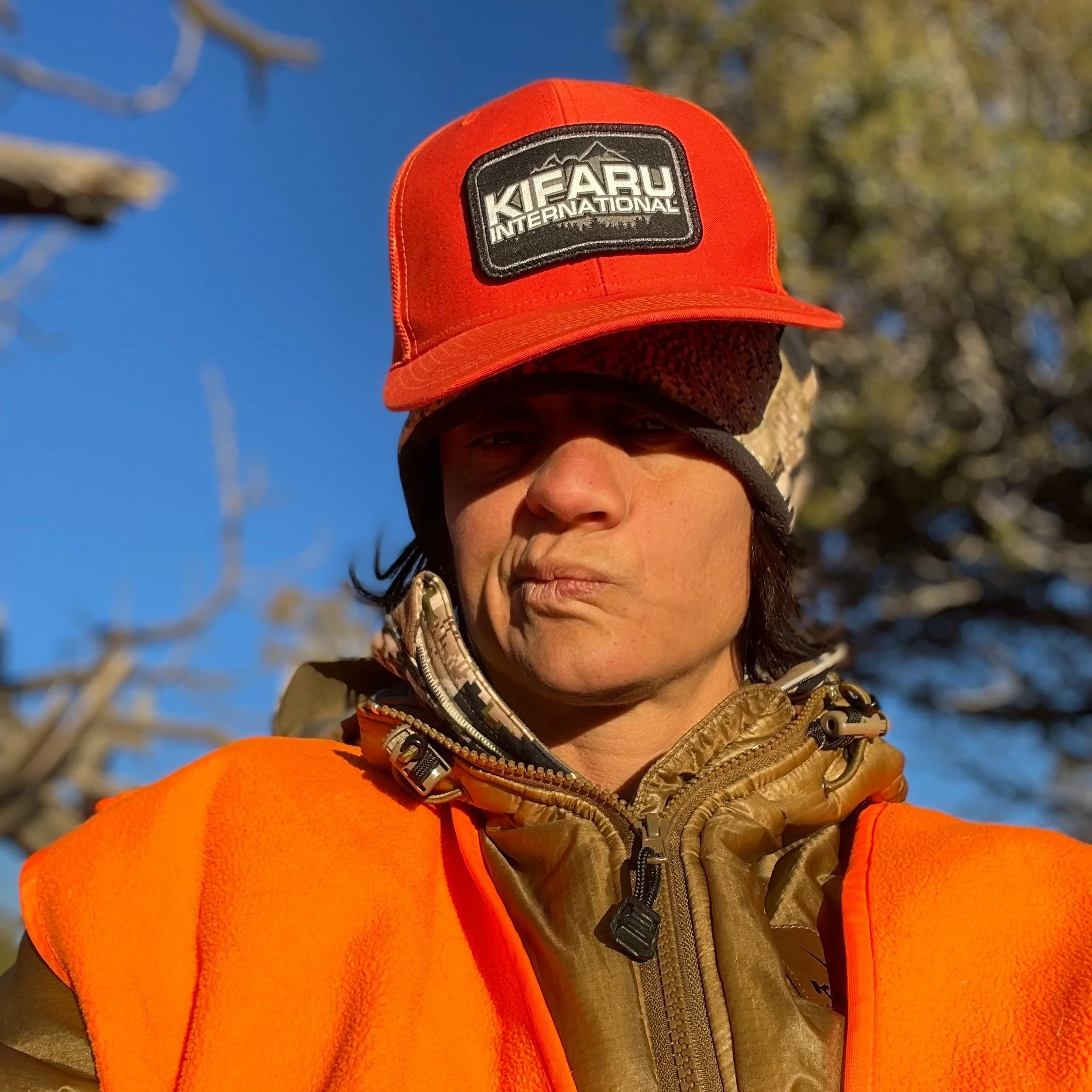 Person wearing a red Kifaru International cap, a brown jacket, and an orange vest outdoors with a clear blue sky and trees in the background.