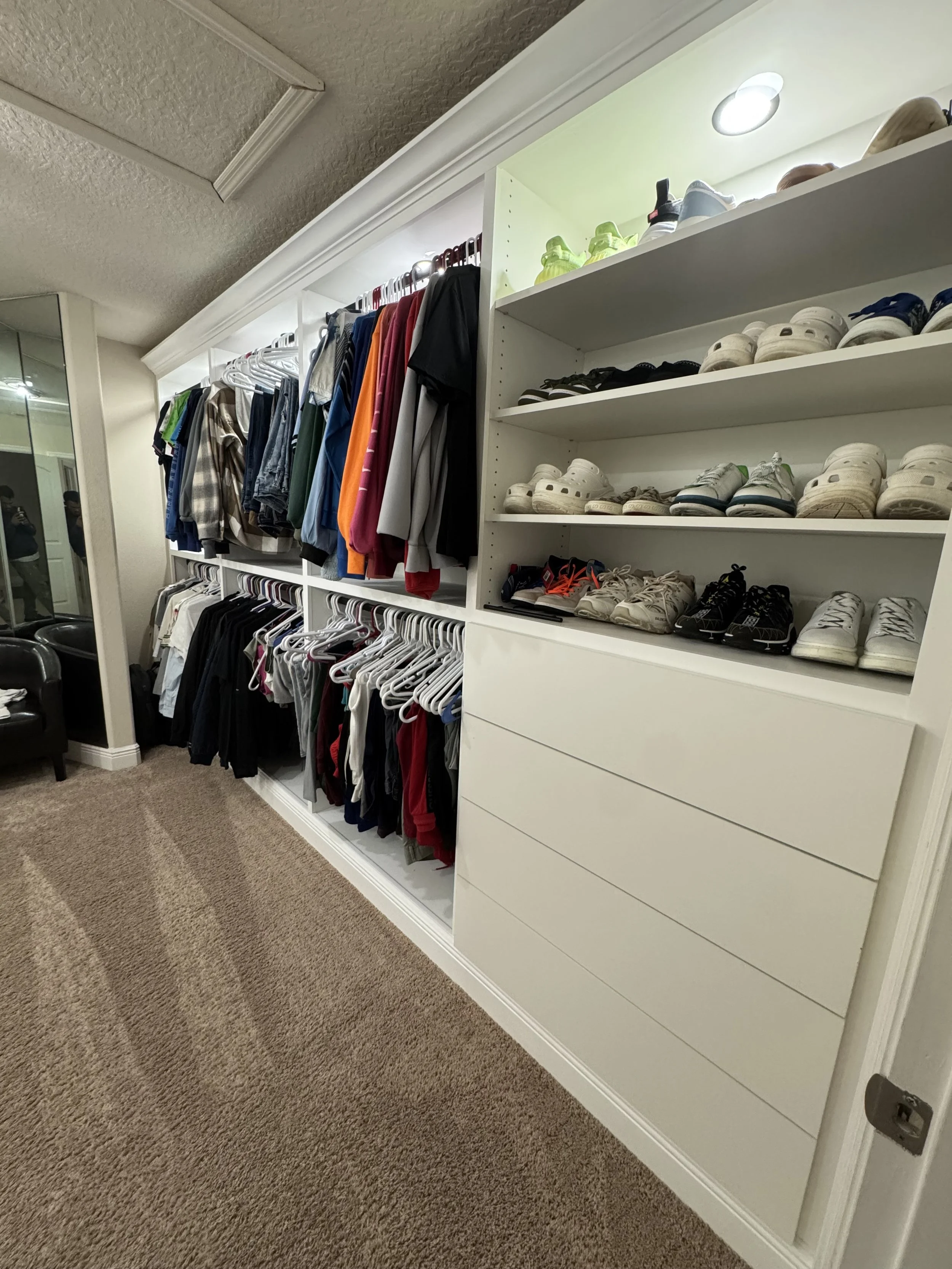 Closet with scrupulously organized shoes on shelves and clothing on hanging racks, beige carpeted floor, and mirrored wall.
