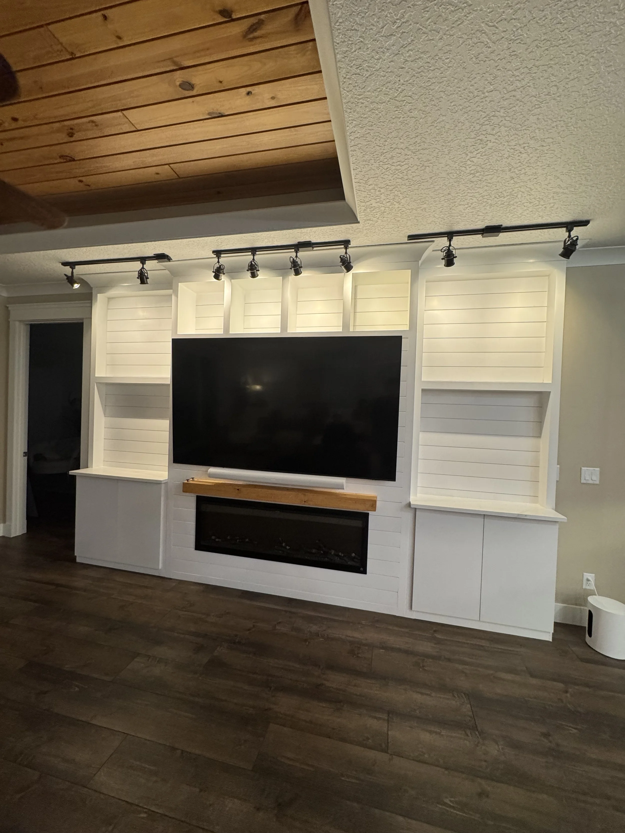Living room with a white built-in entertainment center, a large flat-screen TV, a wooden mantel, and an electric fireplace beneath the TV. Track lighting is installed above, and the room has dark wooden flooring.