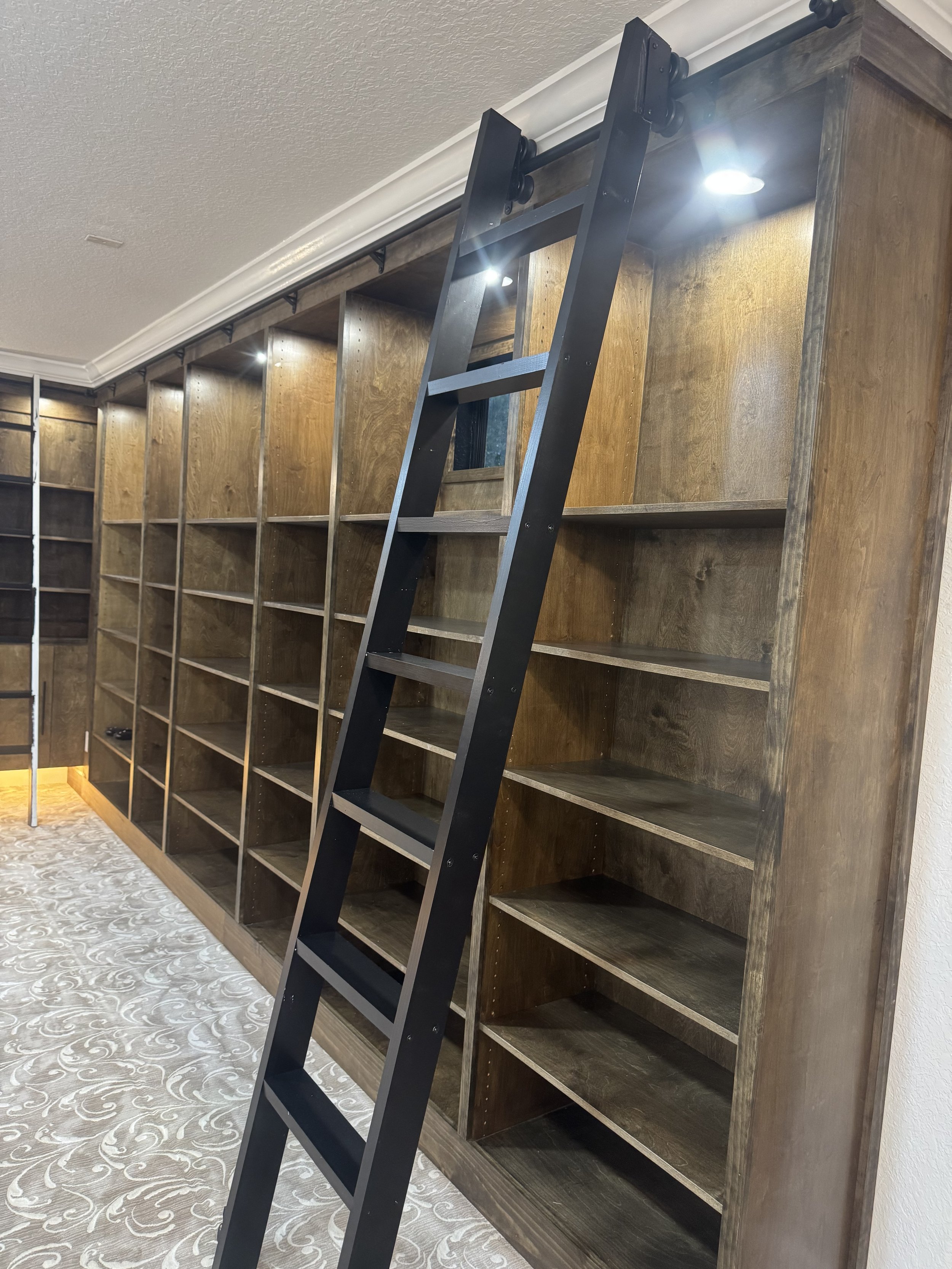 Image of a black step ladder leaning against a wooden bookshelf with multiple empty shelves in a room with beige patterned carpet and ceiling spotlights.