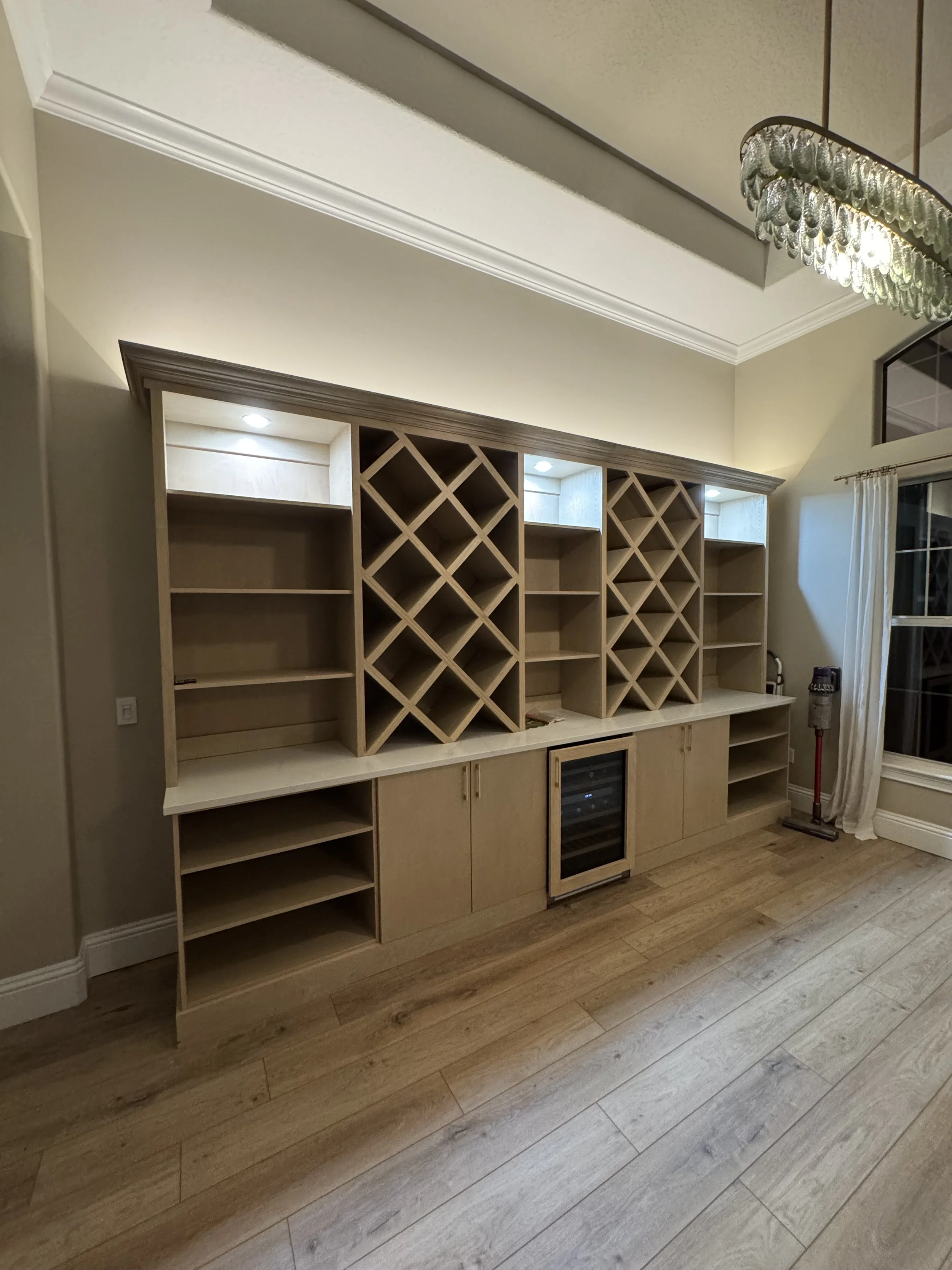 Built-in wooden wine rack and shelving unit in a room with hardwood floor, a wine cooler, and a vacuum cleaner nearby.