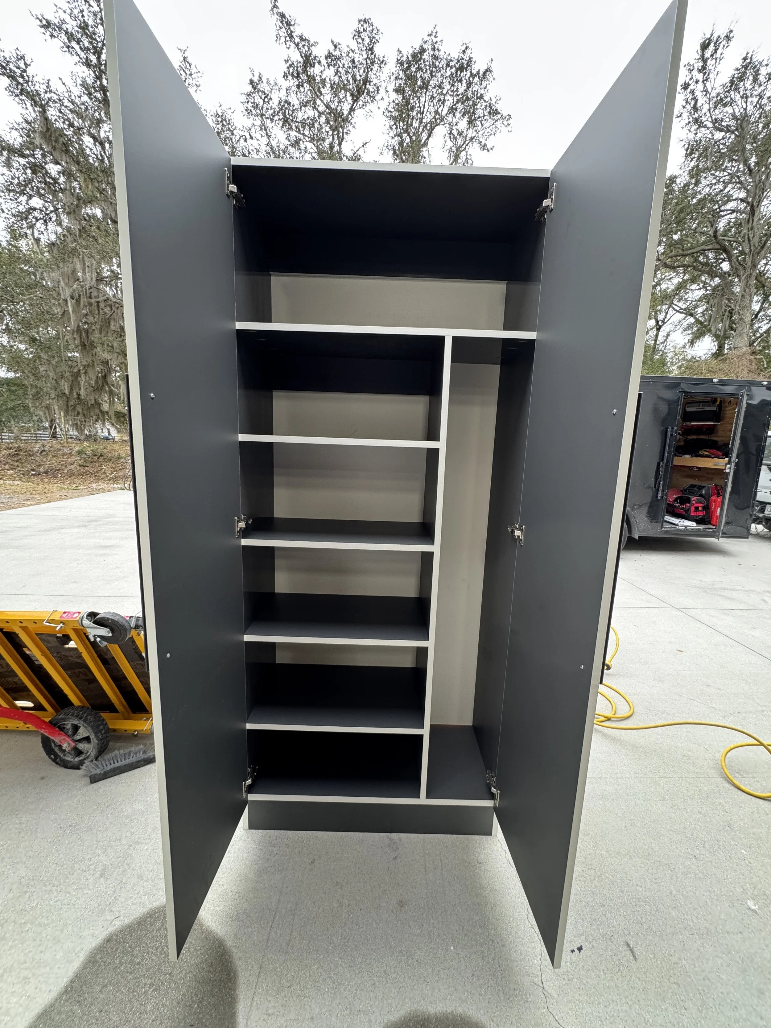 Open black storage cabinet with multiple shelves and doors, standing outdoors on pavement, with trees and a black trailer in the background.