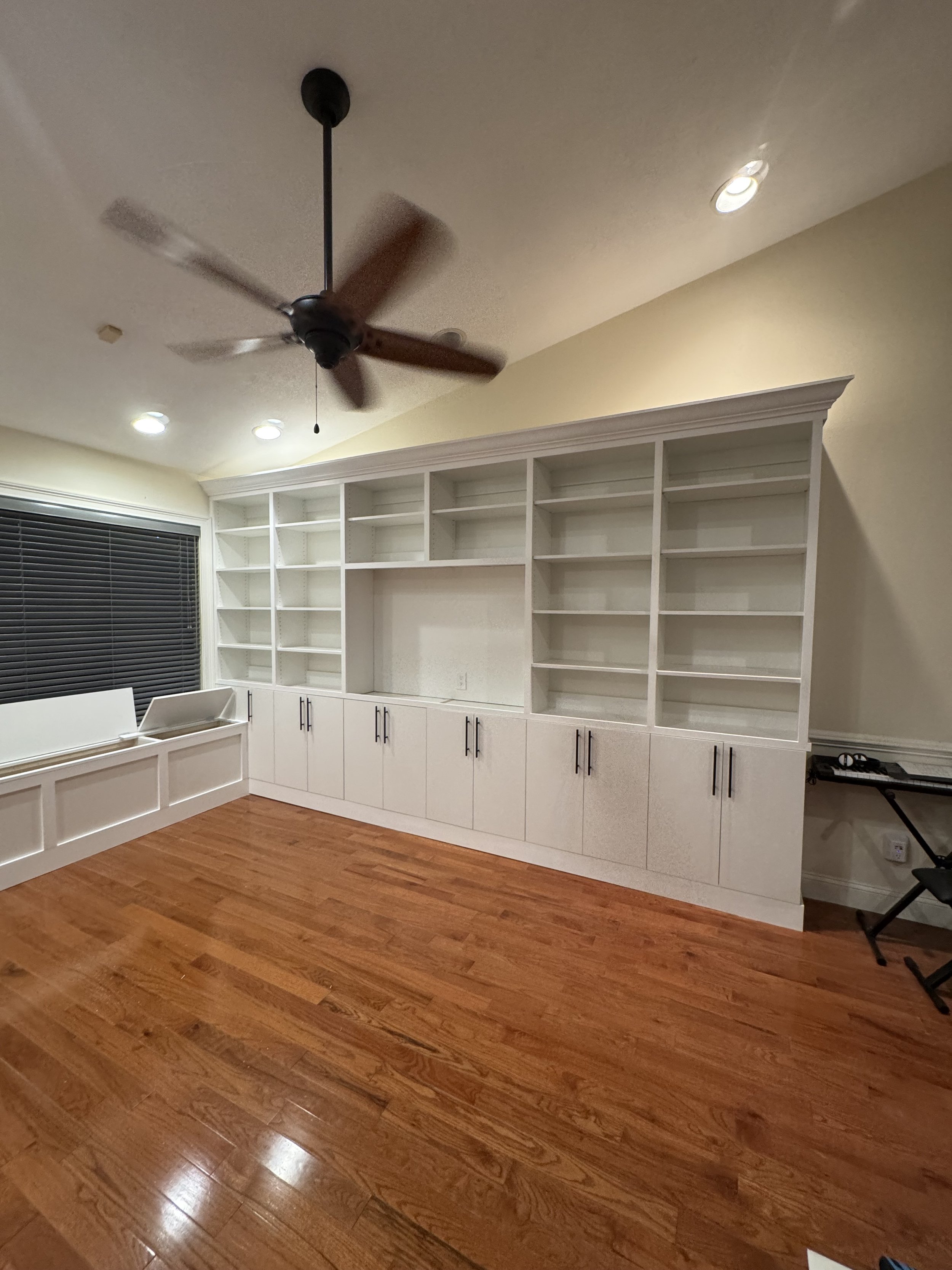 Empty built-in white bookshelves on a wall, hardwood floor, black ceiling fan, window with blinds, and a small black chair in the corner.
