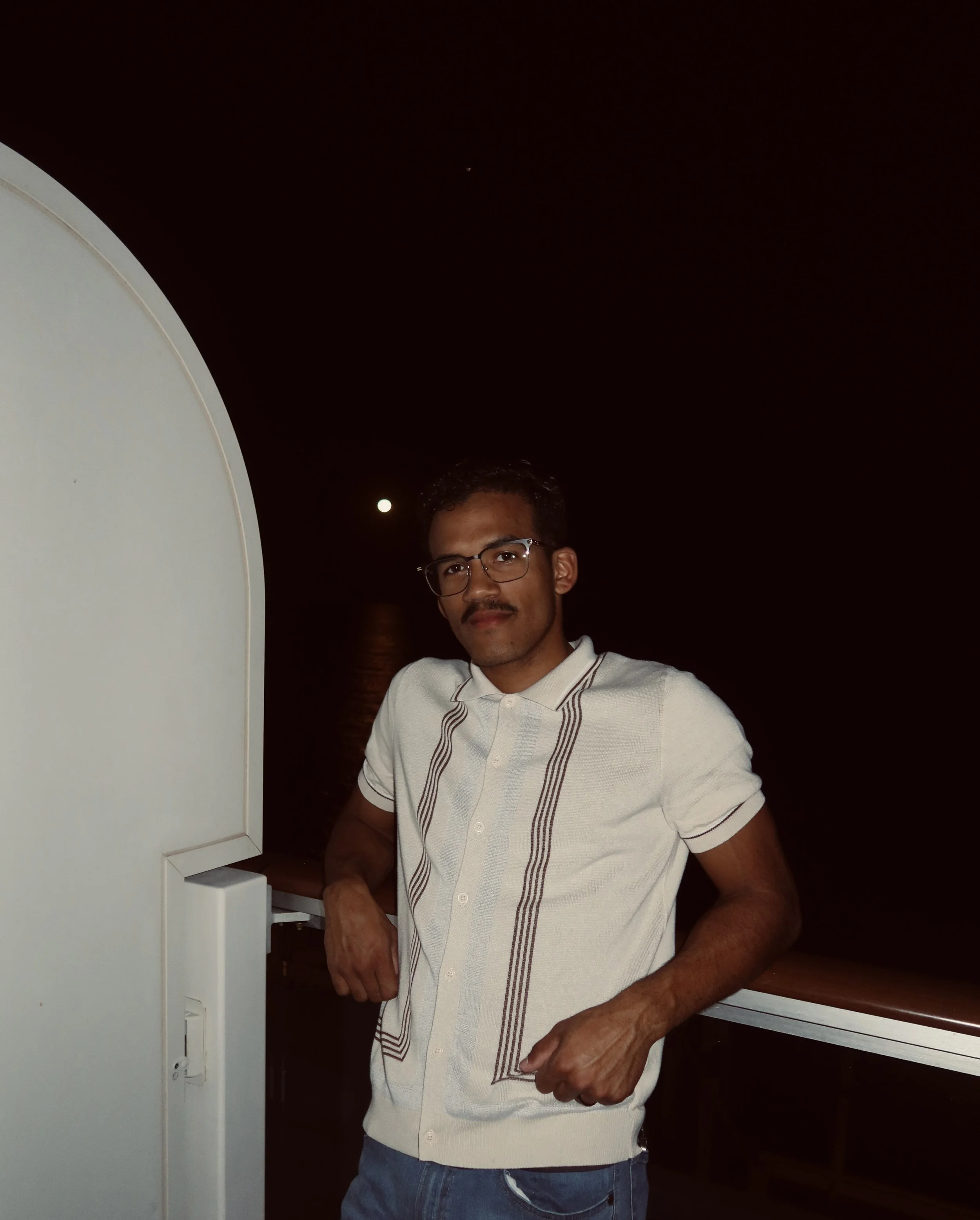 A man with glasses and a mustache standing on a boat at night, with a dark sky and moon in the background.
