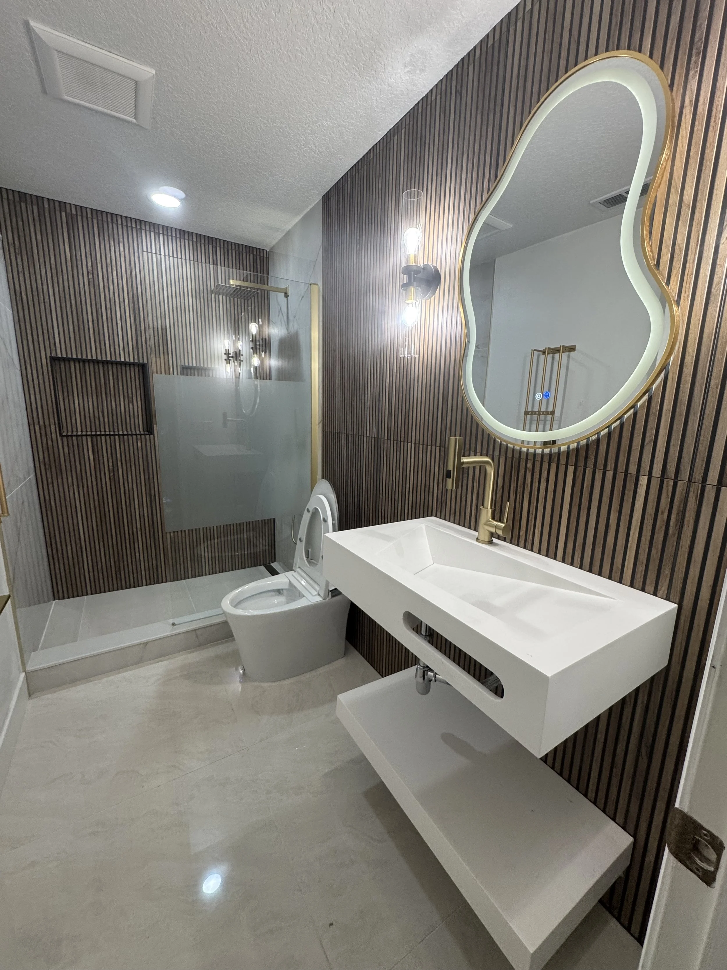 Modern bathroom with a walk-in shower, toilet, white rectangular sink with brass fixtures, large mirror with LED lighting, and wooden panel wall decor.