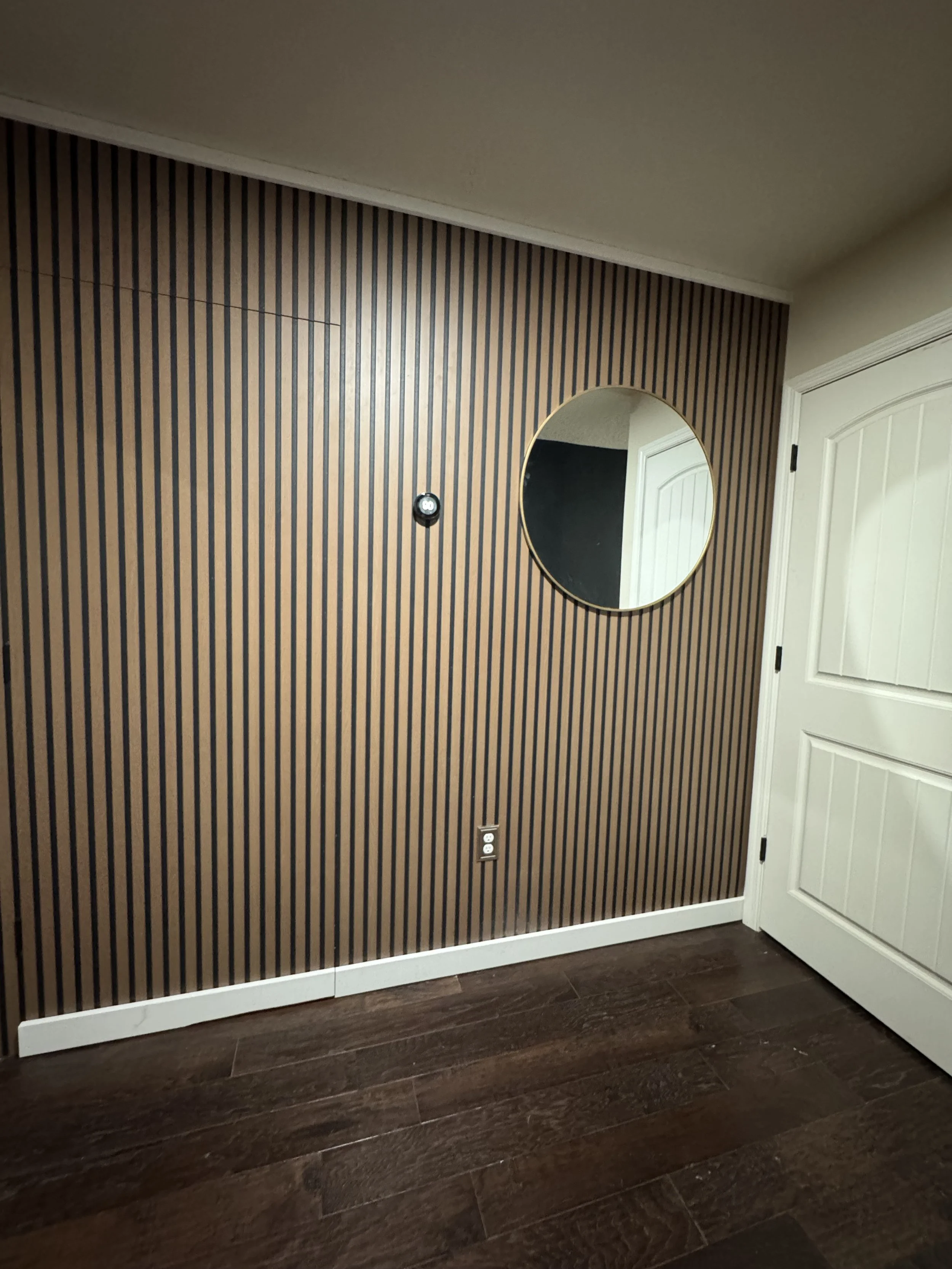 Interior of a room with a wall featuring vertical brown and black striped wallpaper, a round mirror, a white door, and a dark brown wooden floor.