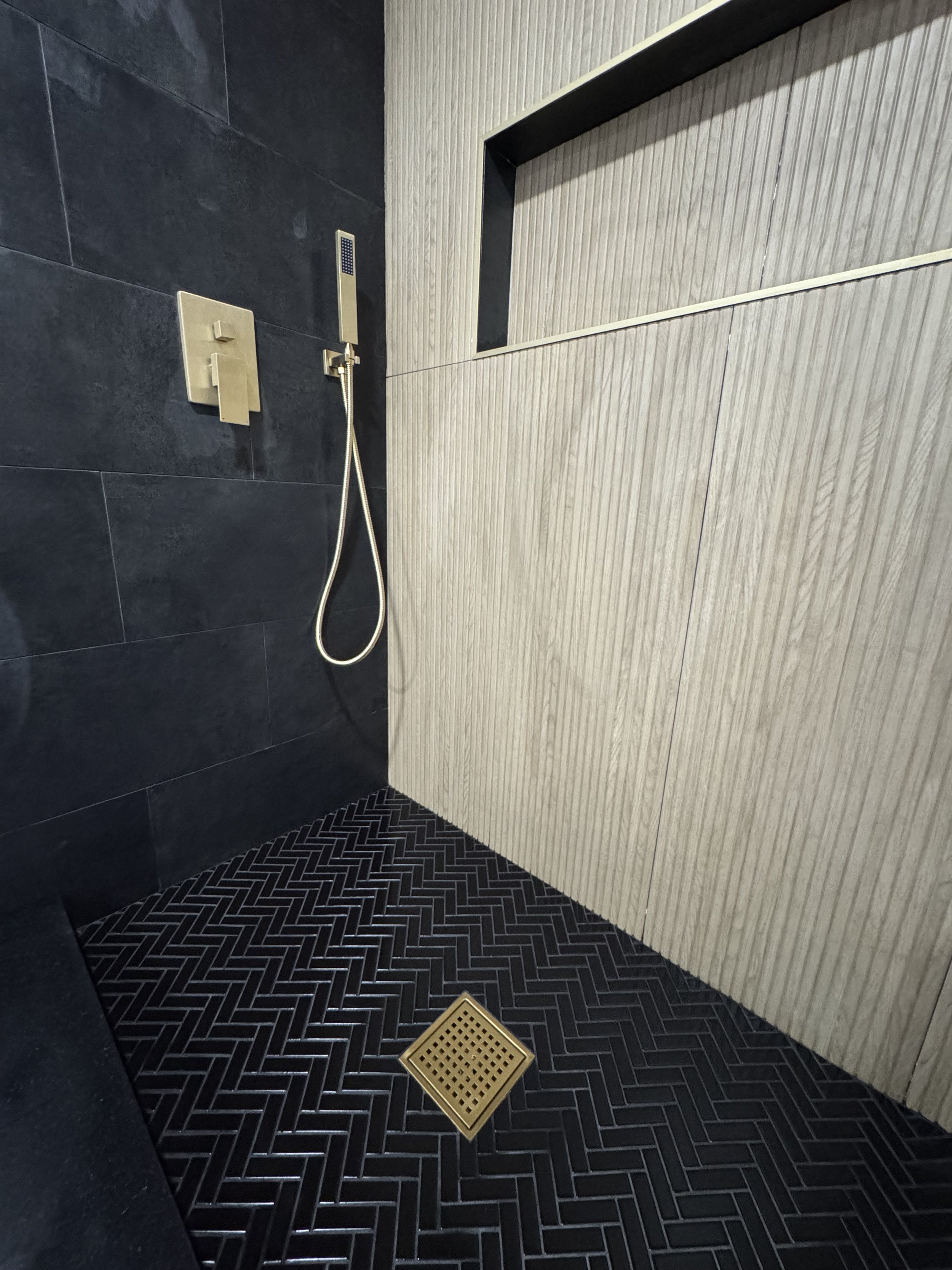 Modern shower space with black tile flooring, a brass drain, a black wall-mounted shower hand wand, and dark gray and light beige textured wall tiles.