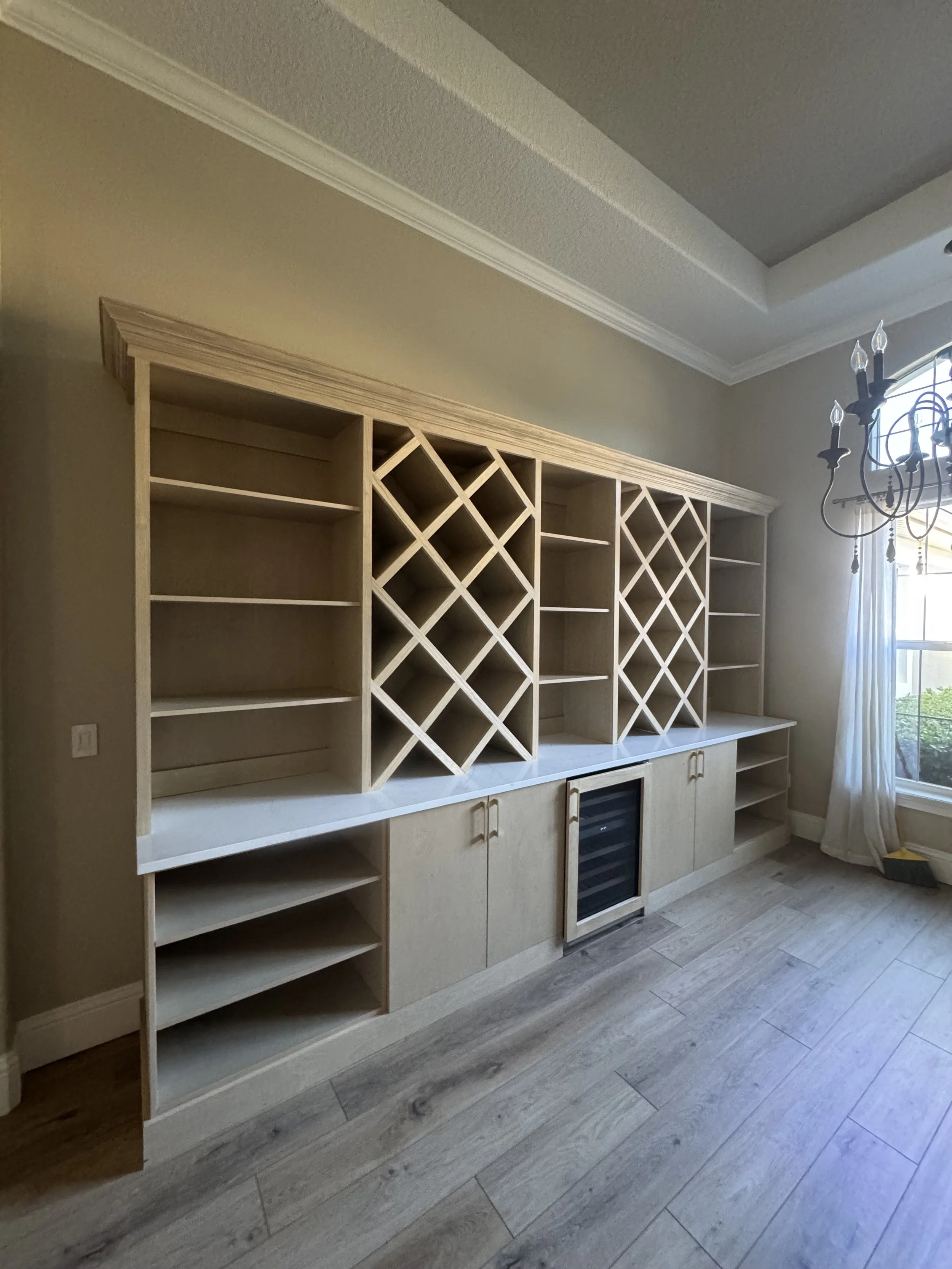 Empty wooden built-in wine and storage cabinet in a room with light-colored walls, a window with white curtains, and a chandelier.