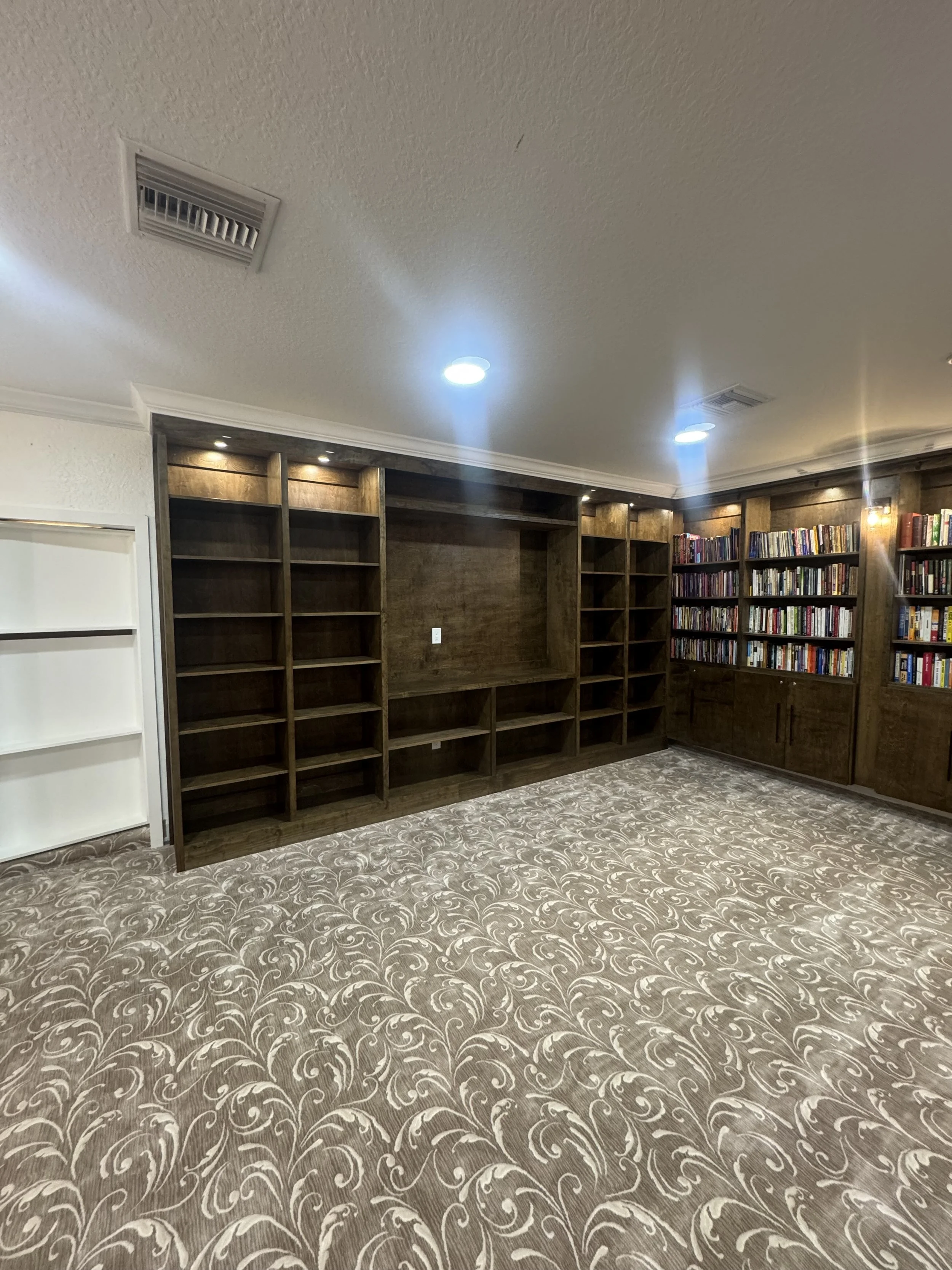 Empty built-in wooden bookshelf with multiple shelves and a nook in the middle, next to a section of closed cabinet with glass doors filled with books, in a room with patterned carpet and ceiling lights.