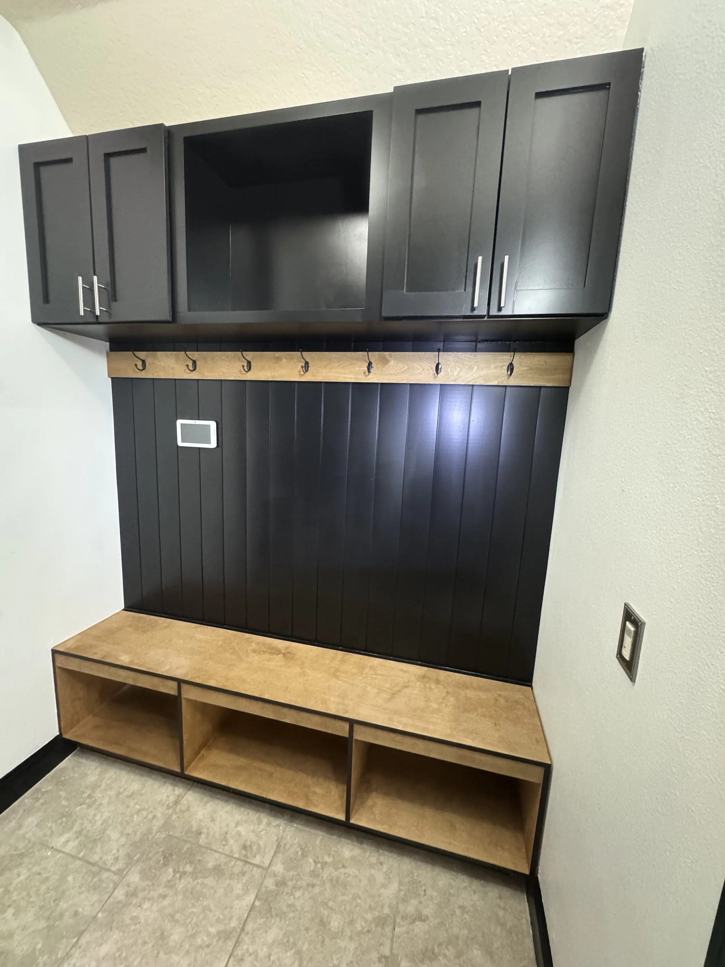 A corner of a room with black upper cabinets, a wooden coat rack with hooks, and a wooden bench with open shelves underneath. The walls are light-colored, and the floor is tiled.