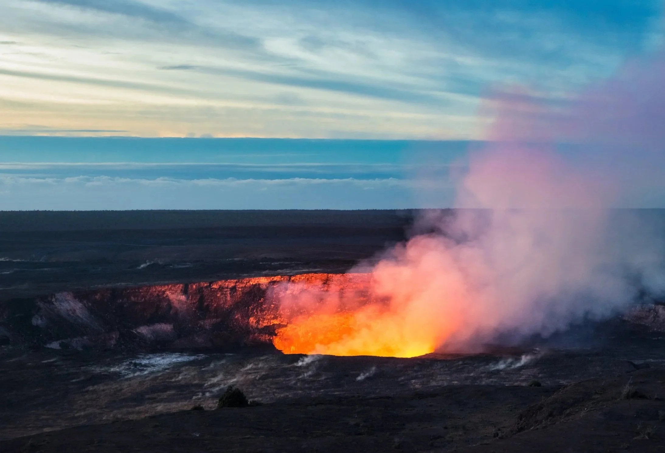 A volcanic eruption with flowing lava, smoke, and ash in the background under a partly cloudy sky.