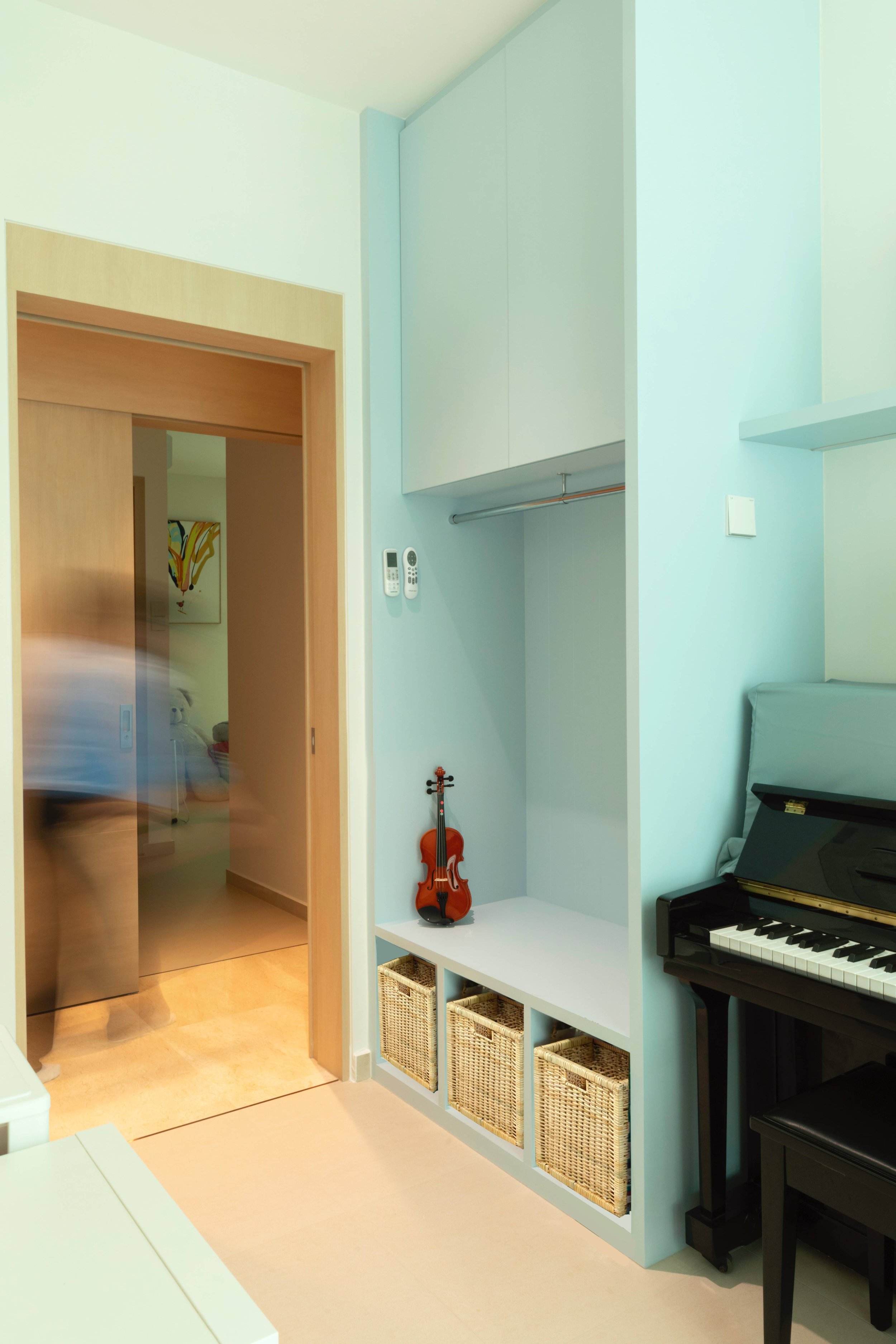 Interior of a room with a light-colored wall and a built-in white shelving unit with baskets. A small orange guitar is placed on the bottom shelf, and a black piano is on the right side. A doorway reveals another room with a painting on the wall.