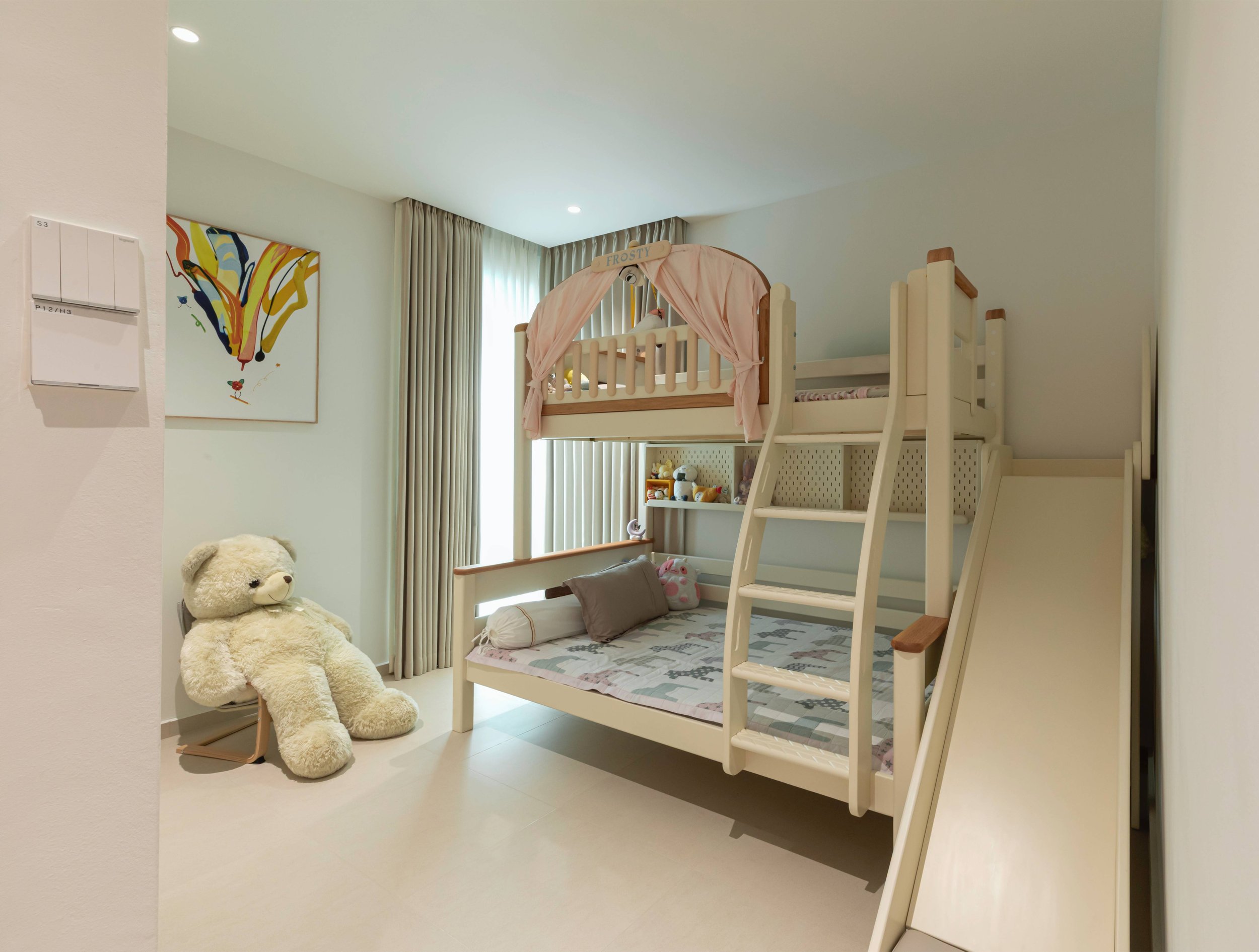 A children's bedroom with a loft bed, a teddy bear sitting on a chair, a colorful abstract painting on the wall, and large windows with beige curtains.