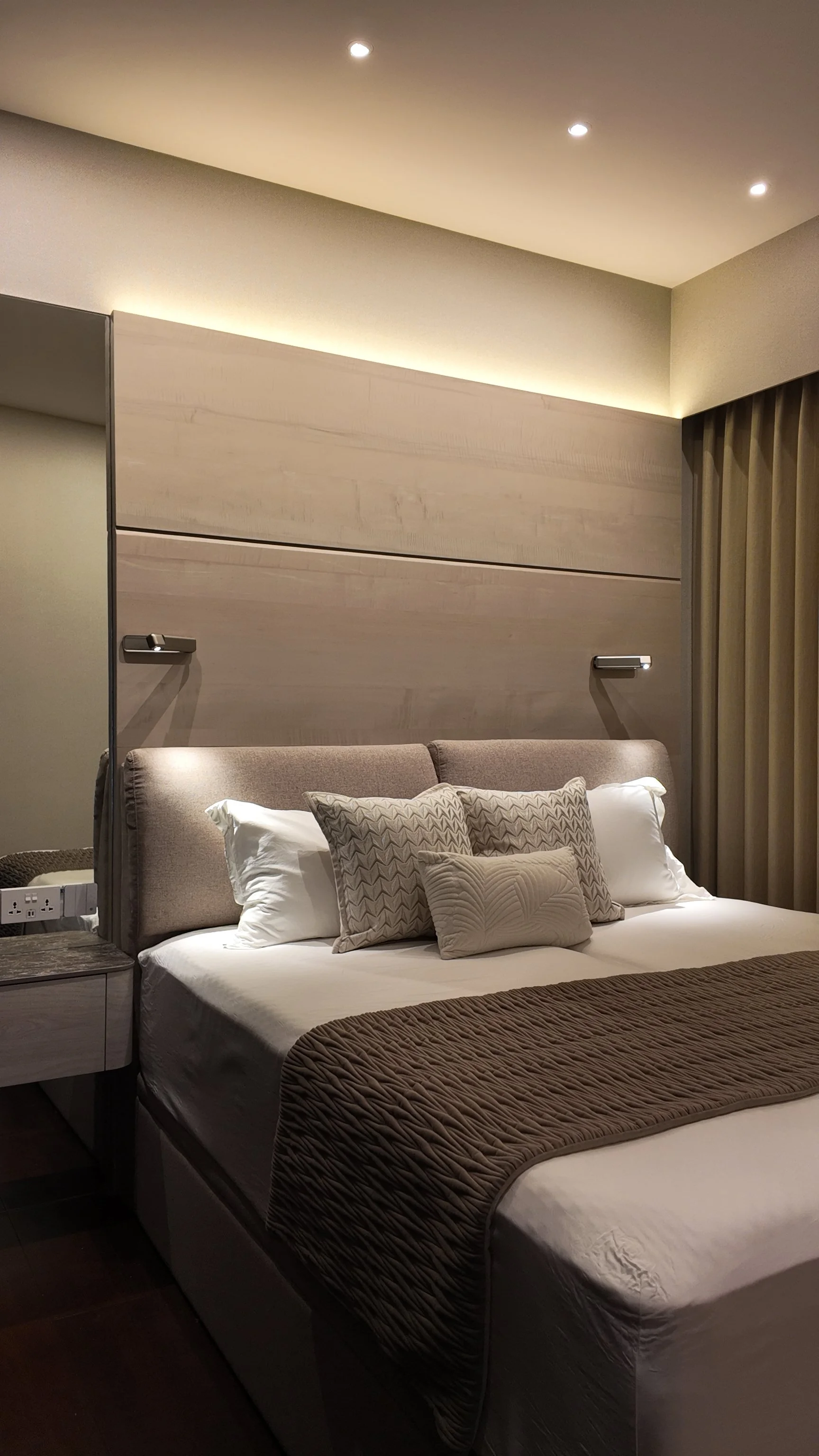 A bedroom with a neatly made bed, beige upholstered headboard, multiple pillows with patterned pillowcases, brown quilted bedspread, bedside table with power outlets, beige curtains, and warm overhead lighting.