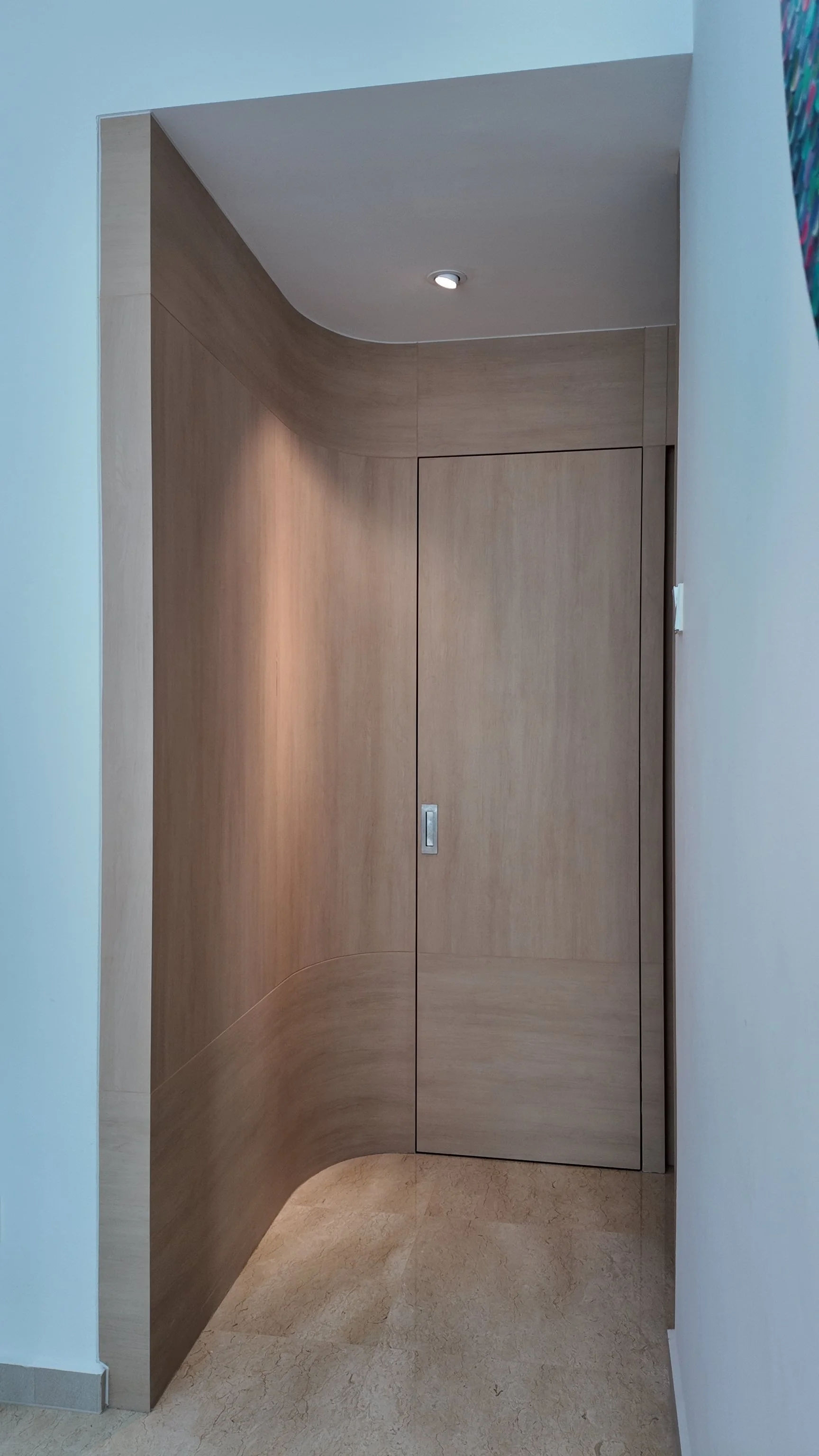 A beige wooden curved corner with a built-in cabinet behind a closed sliding door, set against a light blue wall, with a ceiling light fixture shining down.