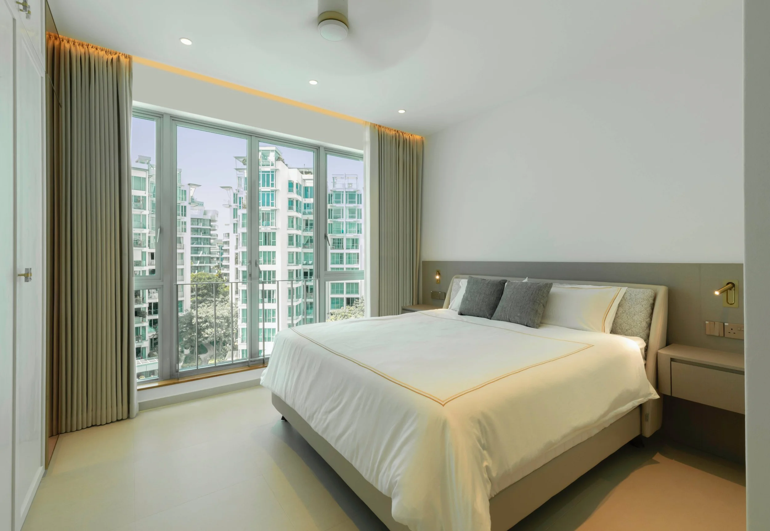 Modern bedroom with large window overlooking tall city buildings, light-colored walls, bed with white linens and gray pillows, beige headboard, nightstands, and built-in closet with sliding doors.