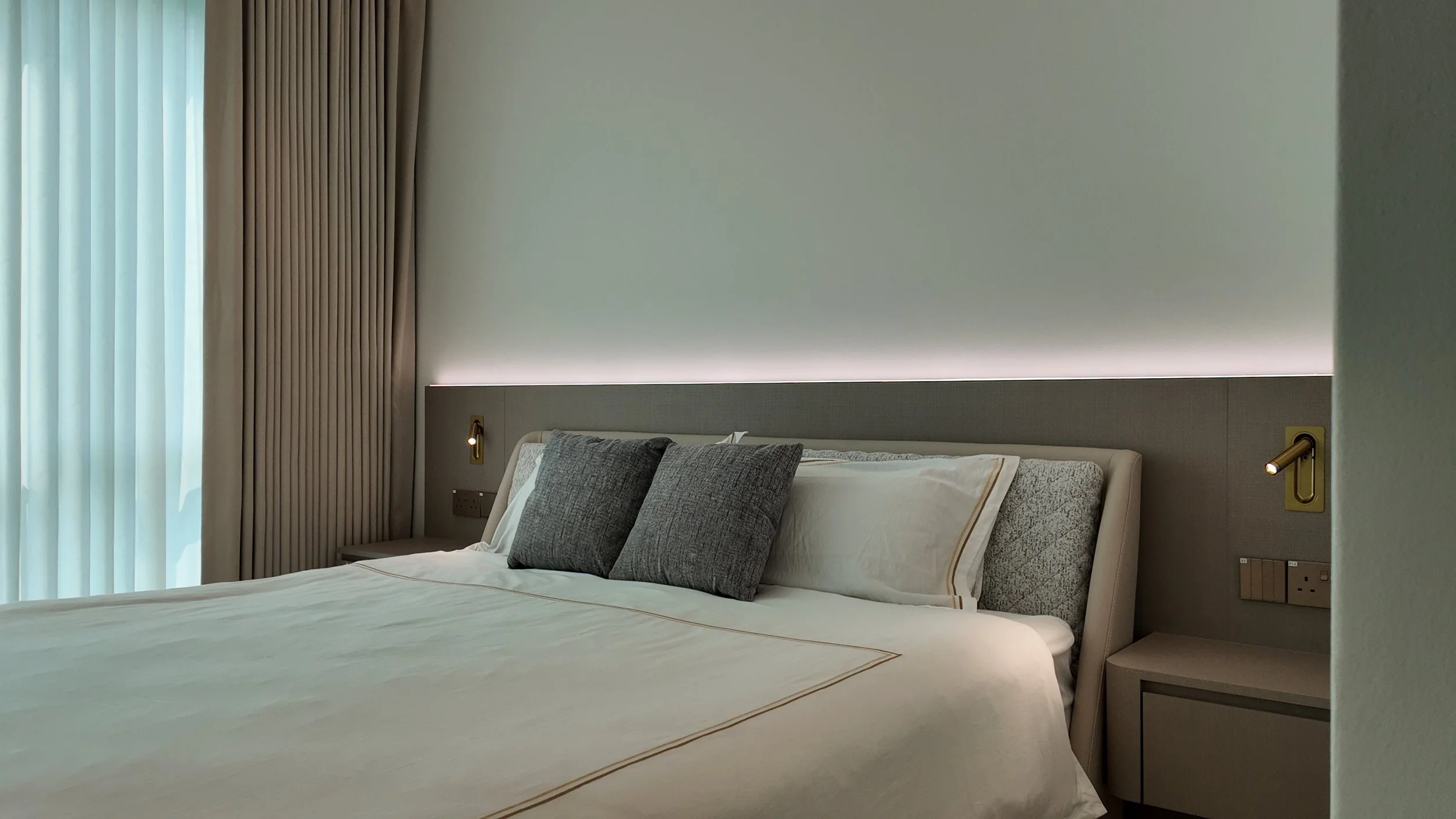 A neatly made bed with white linens, two dark gray pillows, and a light-colored headboard in a modern hotel room. There are bedside lamps on each side, and light filtering through sheer curtains and beige drapes.