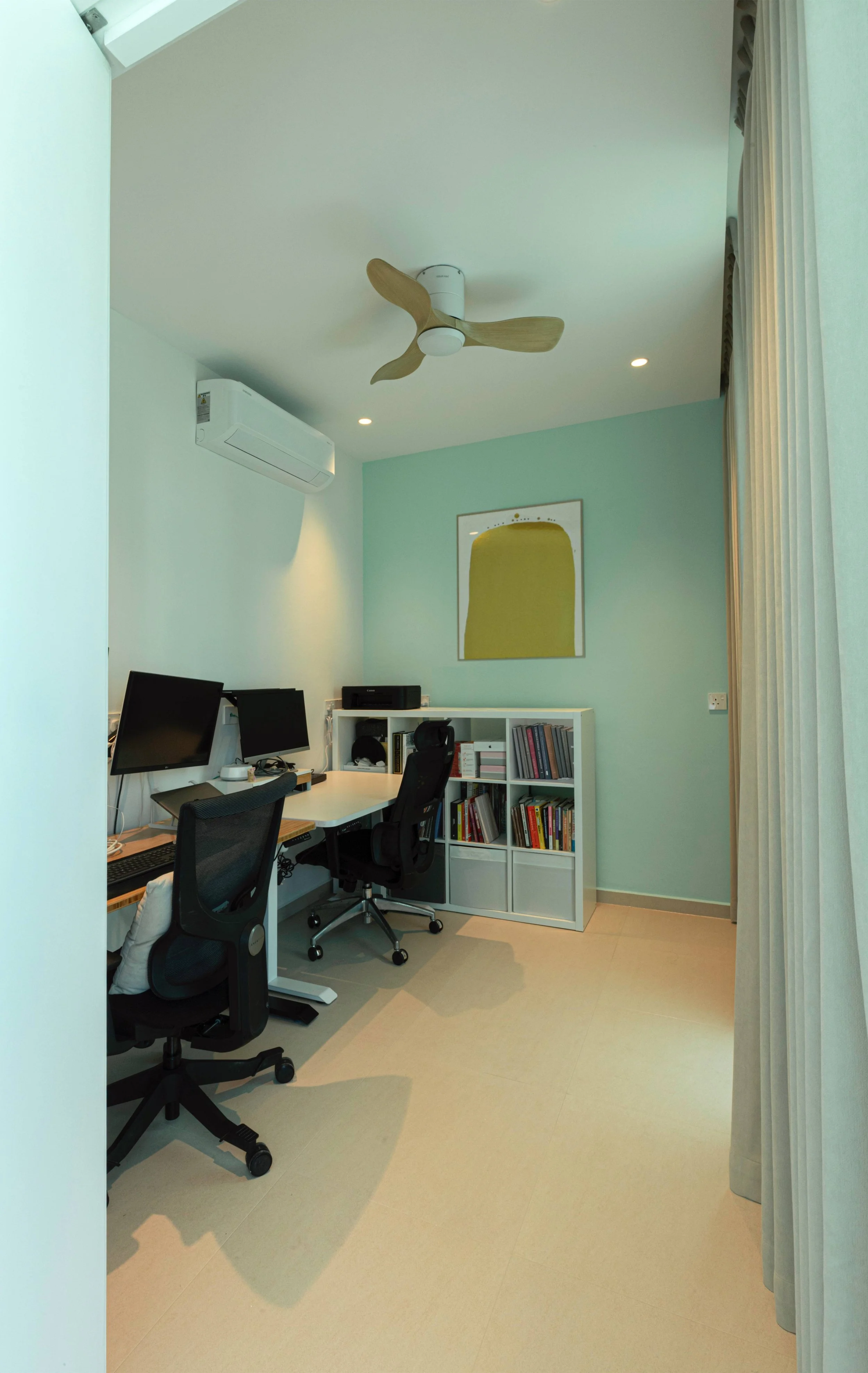 An organized home office with a white desk holding two computer monitors, a black office chair, a white bookshelf filled with books and office supplies, a ceiling fan with wooden blades, a wall-mounted air conditioner, and a piece of abstract artwork on a light green wall next to a large window with white curtains.