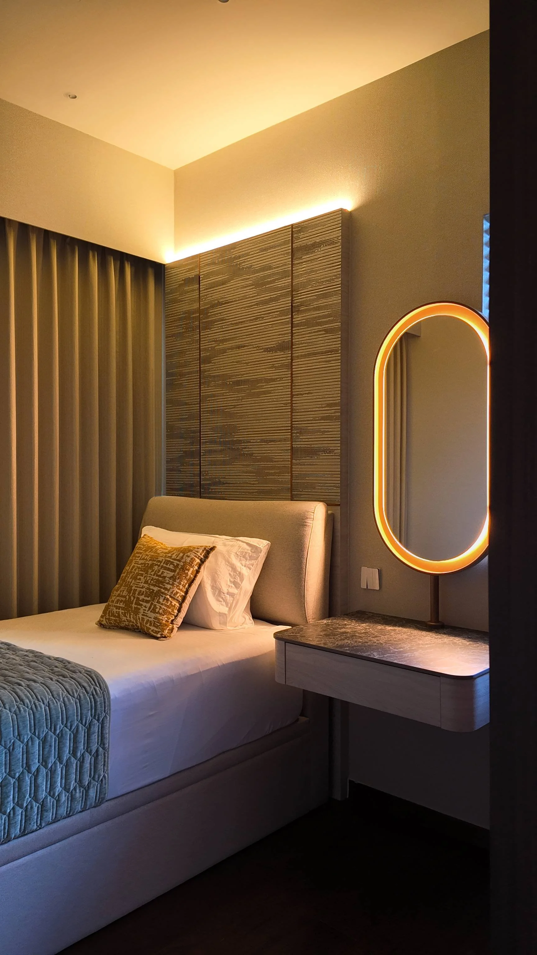 A modern bedroom with a bed, decorative pillows, beige headboard, and a side table with a glowing oval mirror. The room has beige curtains, textured wall panels, and warm lighting.
