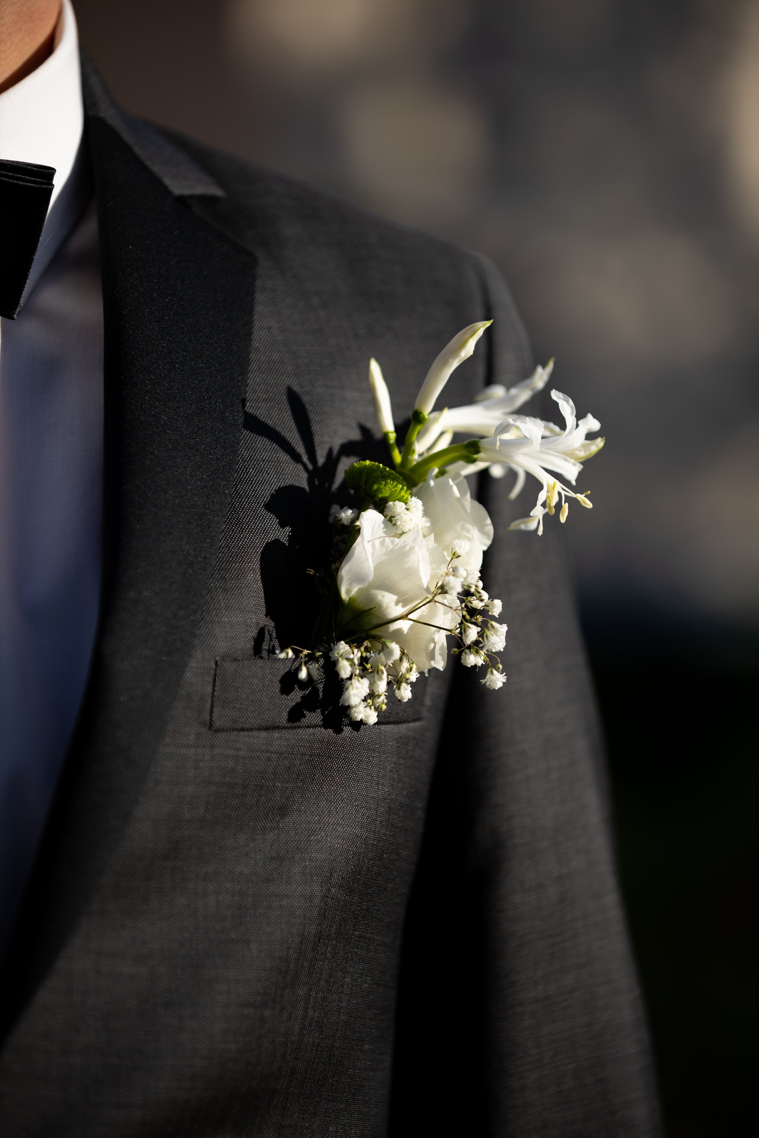 Nahaufnahme eines Anzugs mit einer weißen Blume im Revers. Der Anzug ist dunkelgrau und die Blume besteht aus weißen Blüten und kleinen grünen Blättern.