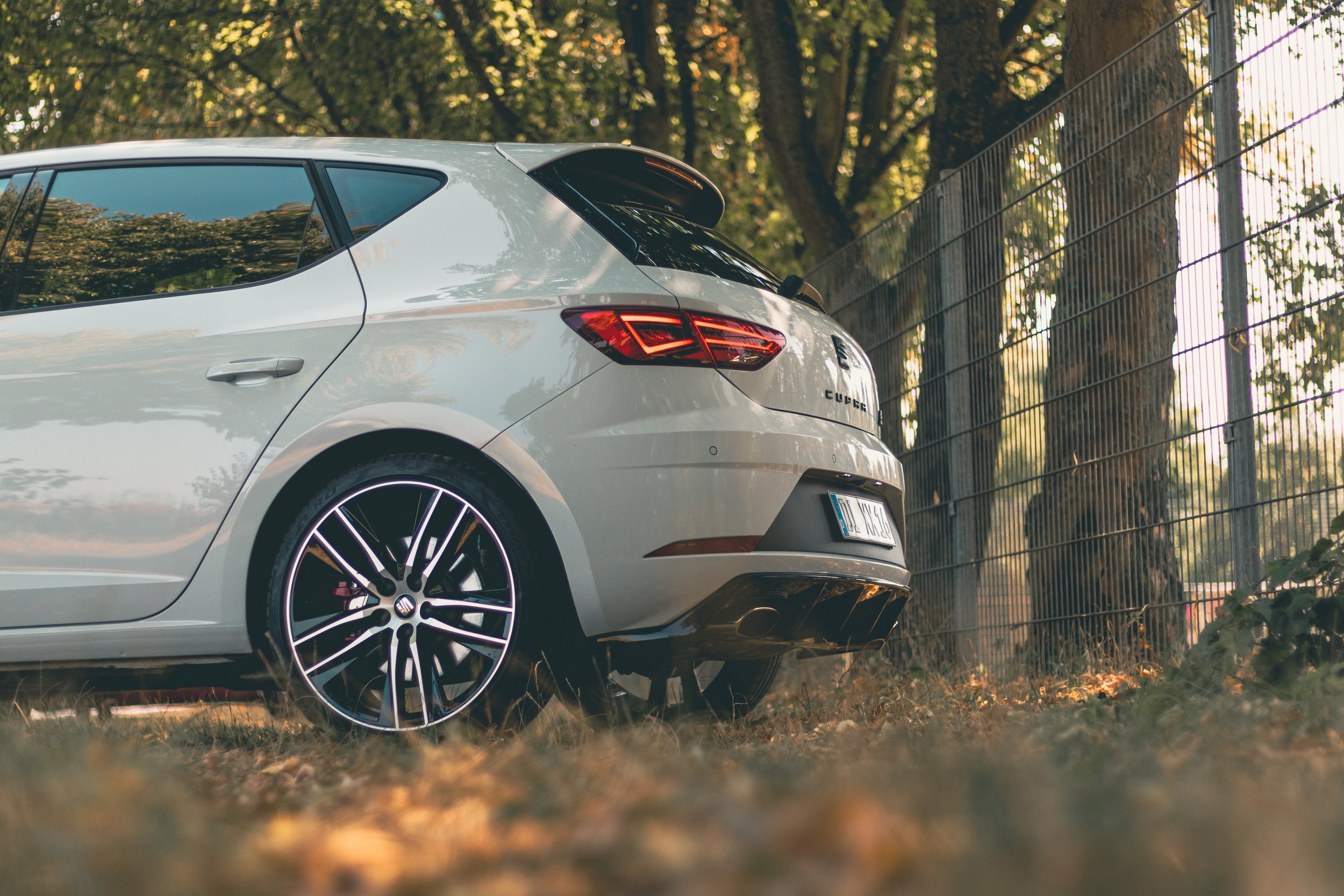 Rückansicht eines weißen Autos mit sportlichem Design, parkiert neben einem Zaun und Bäumen.