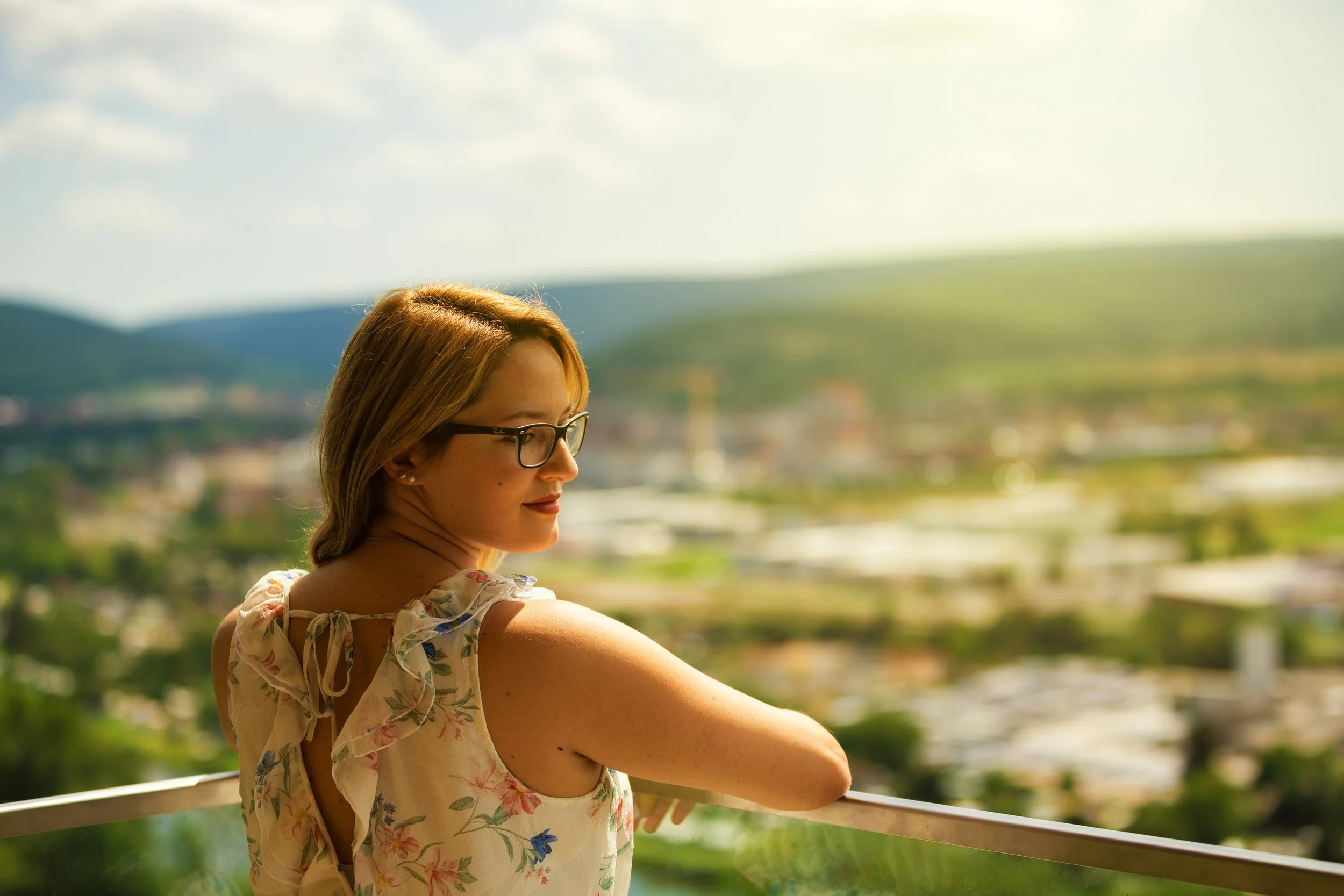 Frau mit Brille und blumigem Kleid blickt von einem Balkon auf eine sonnige Landschaft.