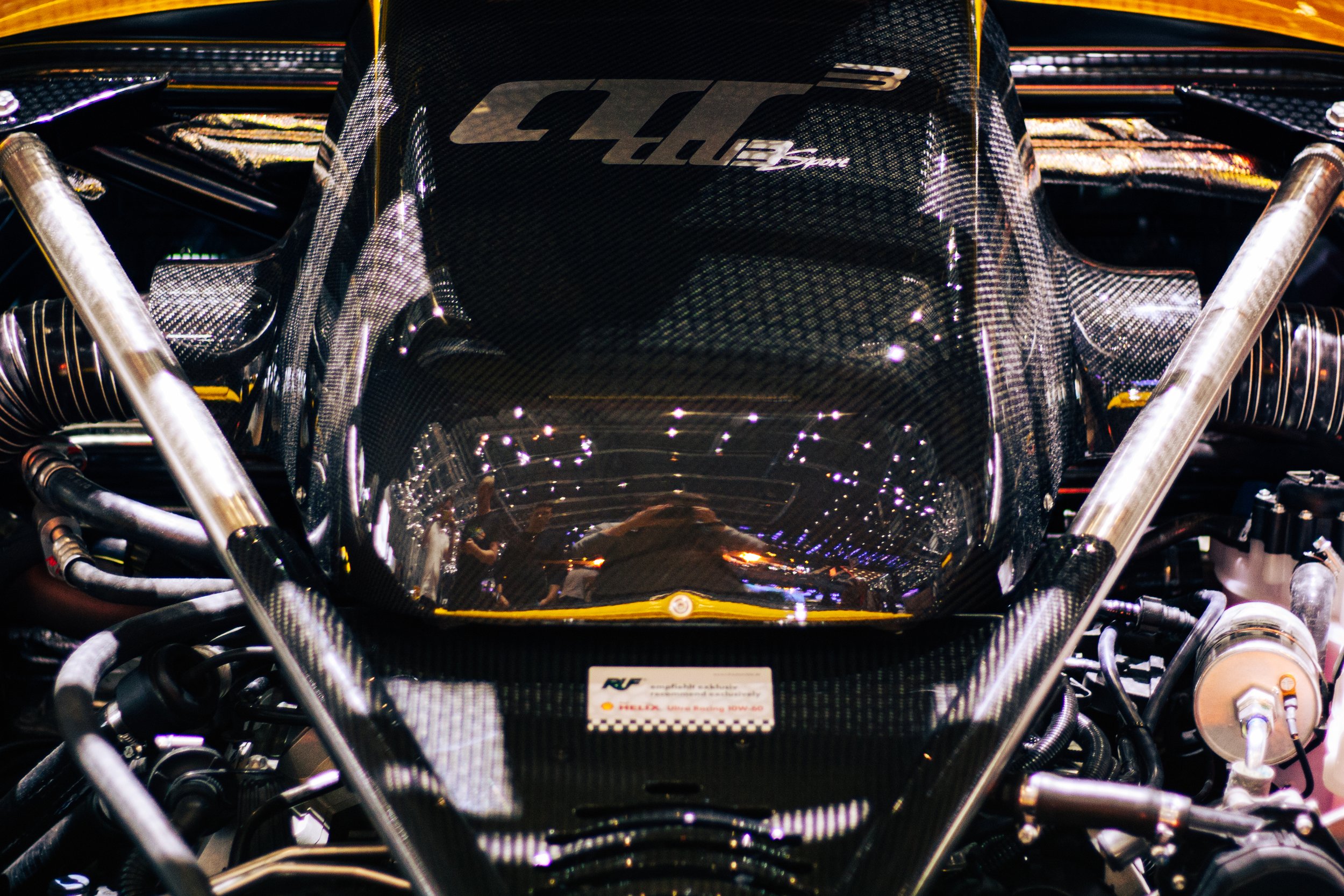 Detailaufnahme eines Autos mit einem Carbon-Motorabdeckung, auf der das Logo "CTR3" zu sehen ist, umgeben von verschiedenen Metall- und Kabelkomponenten.