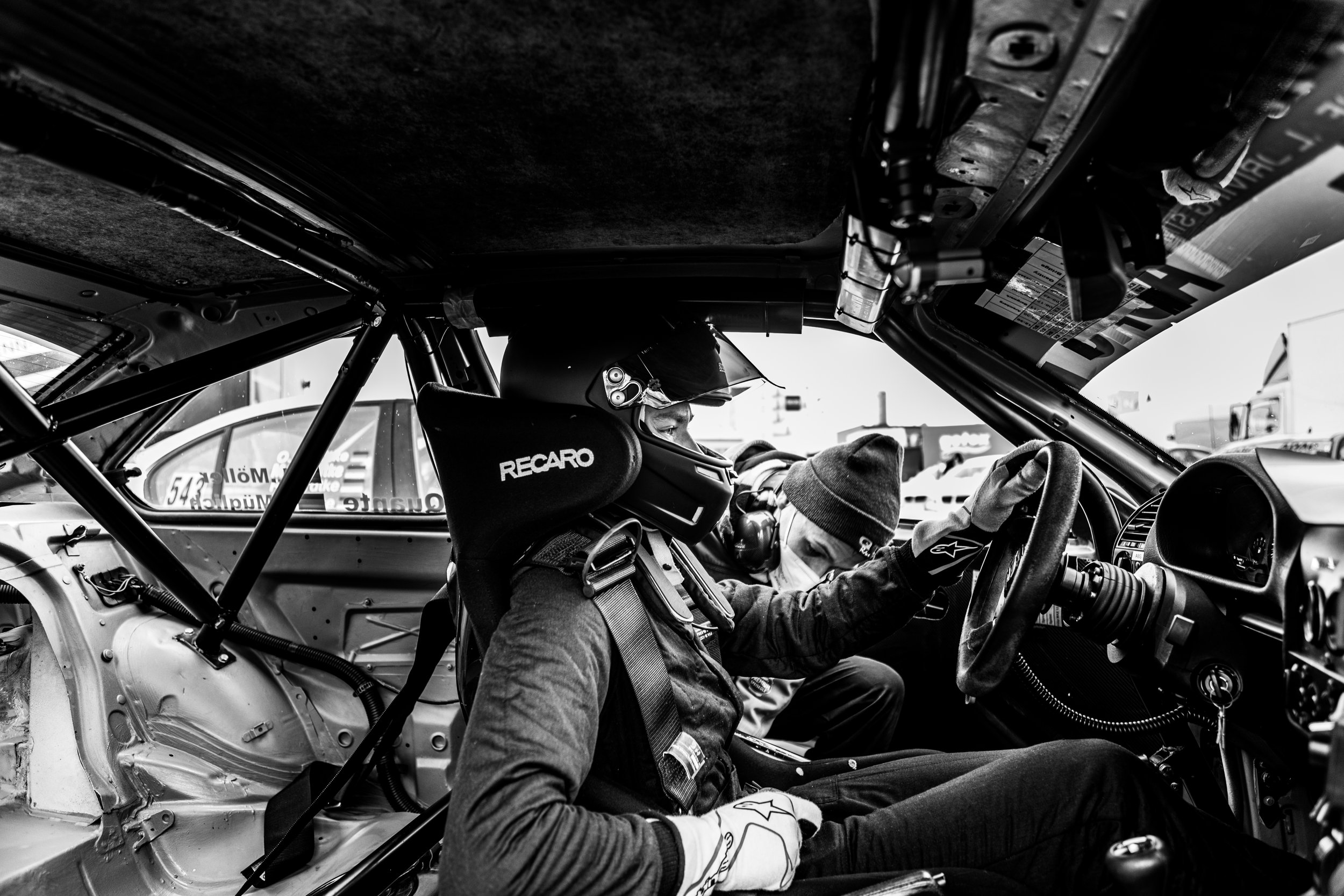 Ein Rennfahrer sitzt in einem Rennwagen mit einem Helm und Sicherheitsgurt. Ein Mechaniker oder Helfer neben dem Fahrer kümmert sich um Einstellungen im Cockpit. Der Innenraum des Wagens zeigt eine komplette Sicherheitsstruktur mit Überrollkäfig.